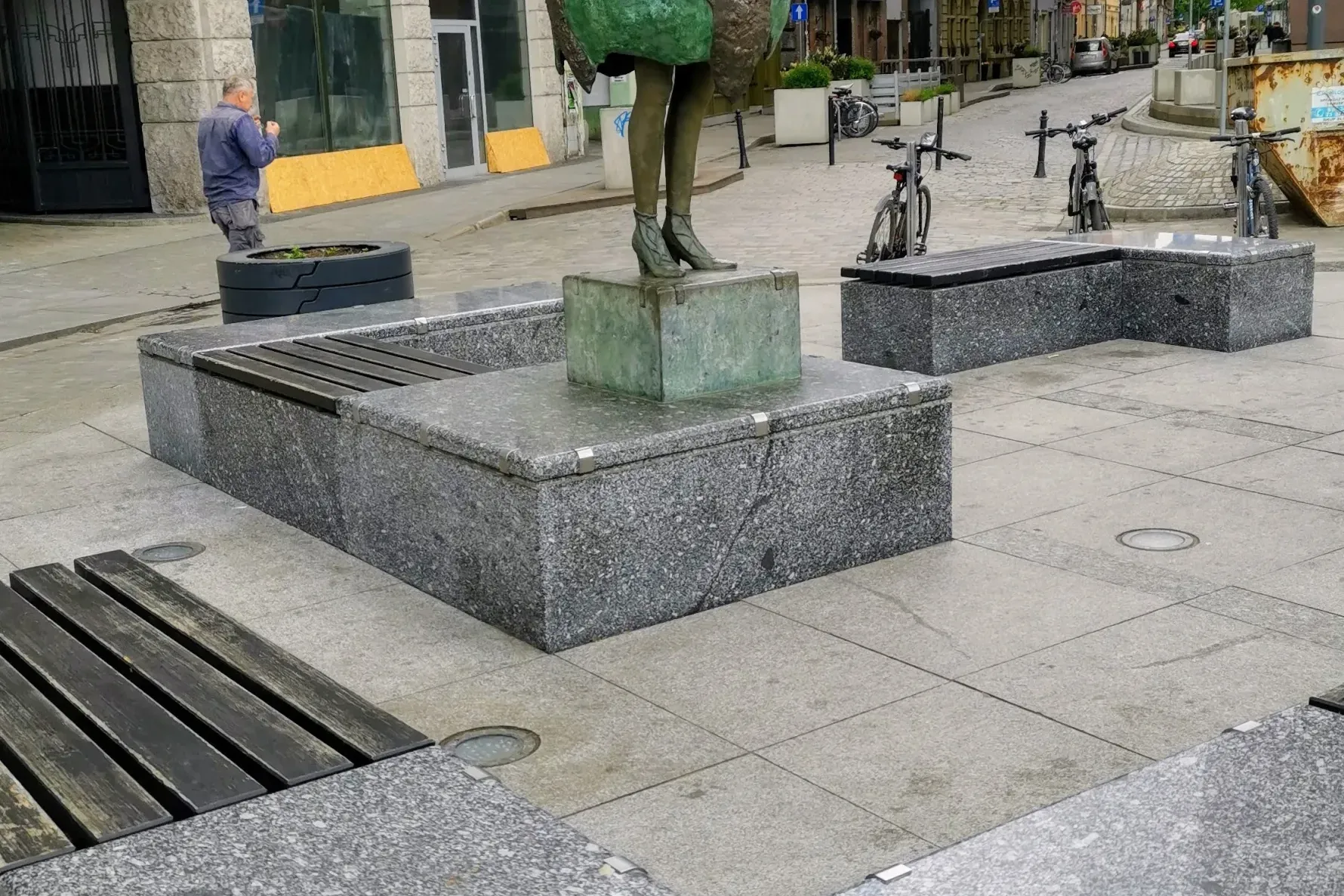 Brązowa statua kobiety z globusem jako spódnicą, granitowe ławki i metalowa tablica z symbolami religijnymi na ulicy.