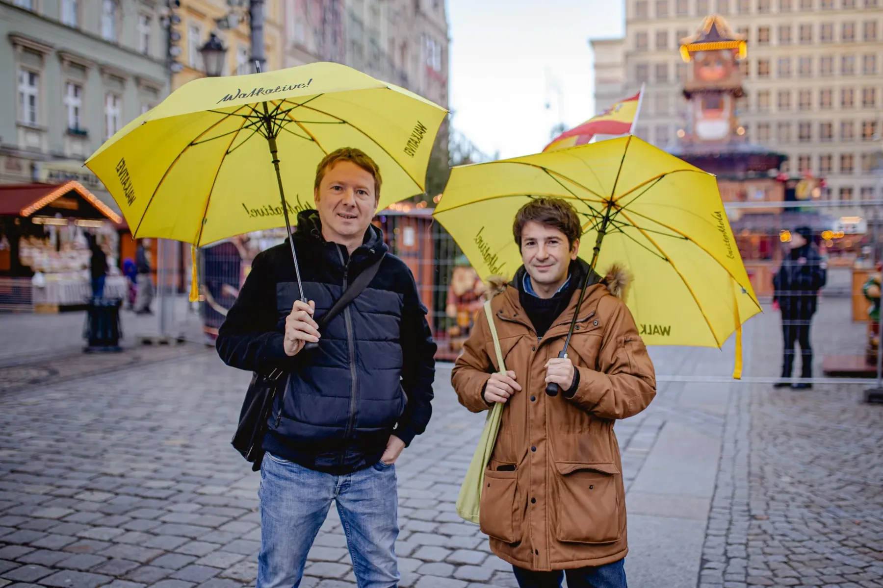 Dwóch mężczyzn z żółtymi parasolami Walkative! stoi na brukowanym placu z niewyraźnymi budynkami i straganami w tle.