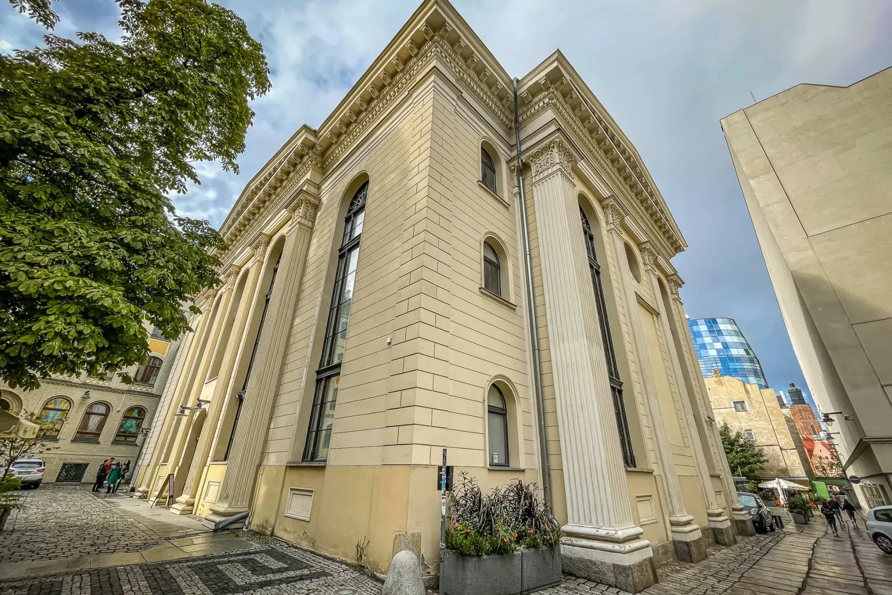 Widok zewnętrzny dużego, jasnożółtego budynku klasycystycznego z kolumnami i łukowymi oknami przy brukowanej ulicy we Wroc...