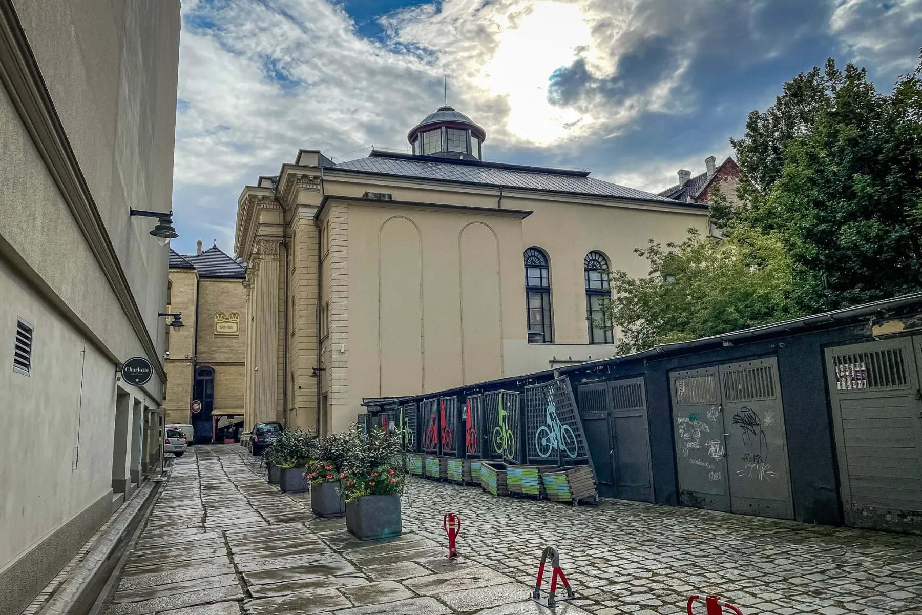 Brukowa uliczka prowadząca do Synagogi Pod Białym Bocianem we Wrocławiu pod zachmurzonym niebem. Widoczne donice i stojaki...