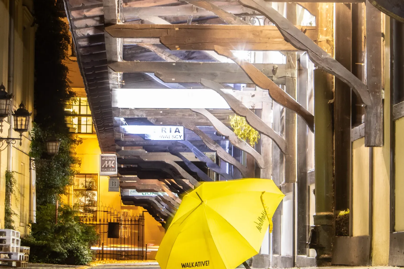Żółty parasol Walkative! na brukowanej ulicy nocą, pod drewnianym pasażem oświetlonym jasnym światłem.
