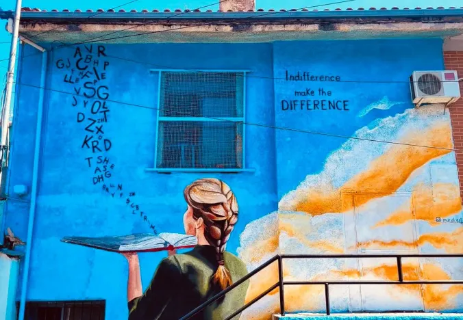 Buntes Street-Art-Wandgemälde einer Frau mit einem Buch an einem blauen Gebäude.