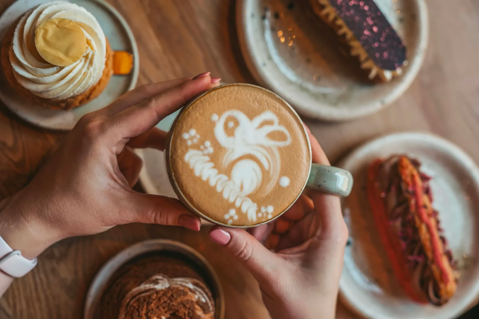Dłonie trzymające filiżankę kawy z latte art, w otoczeniu ciast na drewnianym stole.
