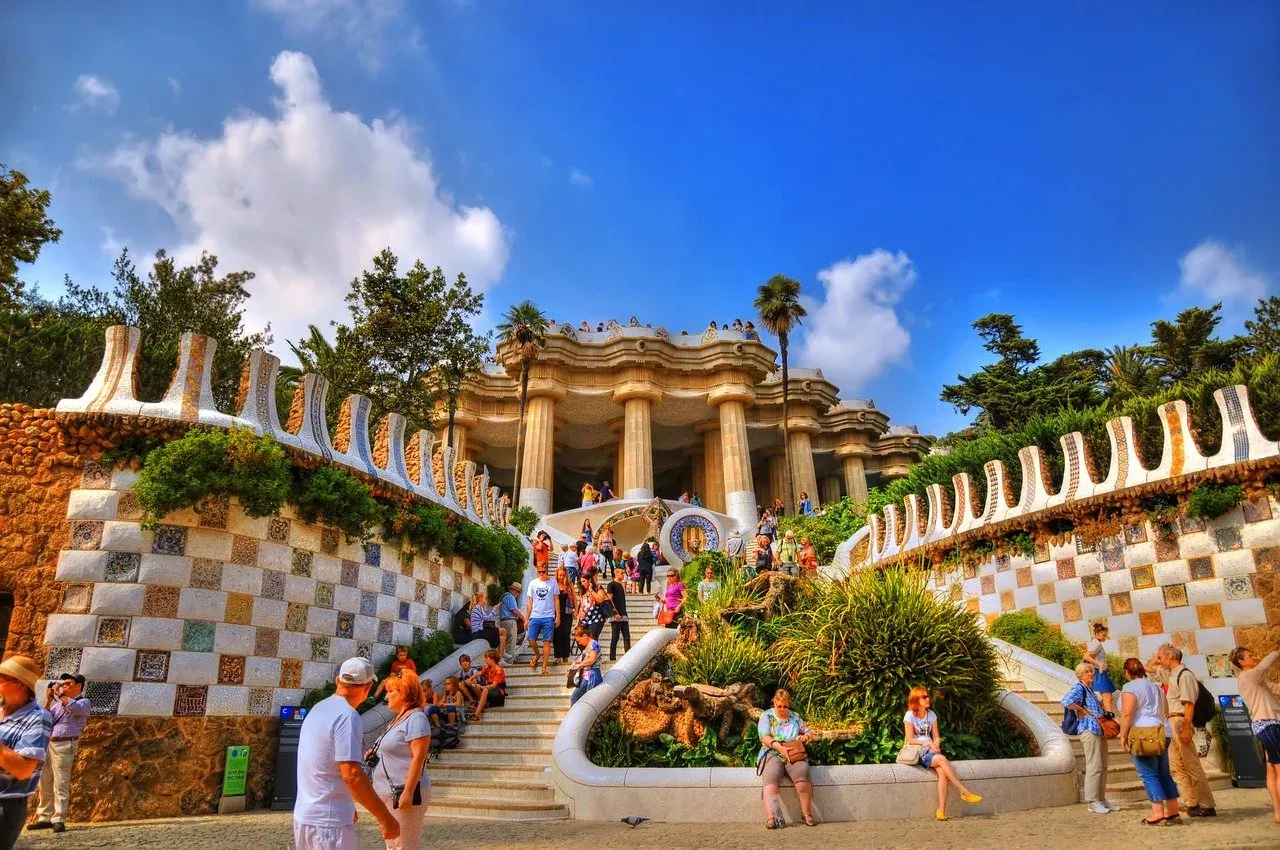 Mosaic-tiled walls and steps lead up to the Hypostyle Room in Park Güell, Barcelona, with numerous tourists on a sunny day.