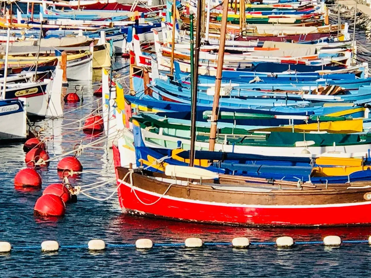Zahlreiche traditionelle, bunte Fischerboote (Pointus) liegen in einem sonnigen Hafen in Nizza, rote Bojen im Vordergrund.