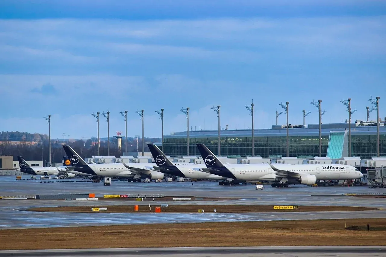 Several Lufthansa passenger airplanes parked at gates at a large airport terminal under a partly cloudy sky.