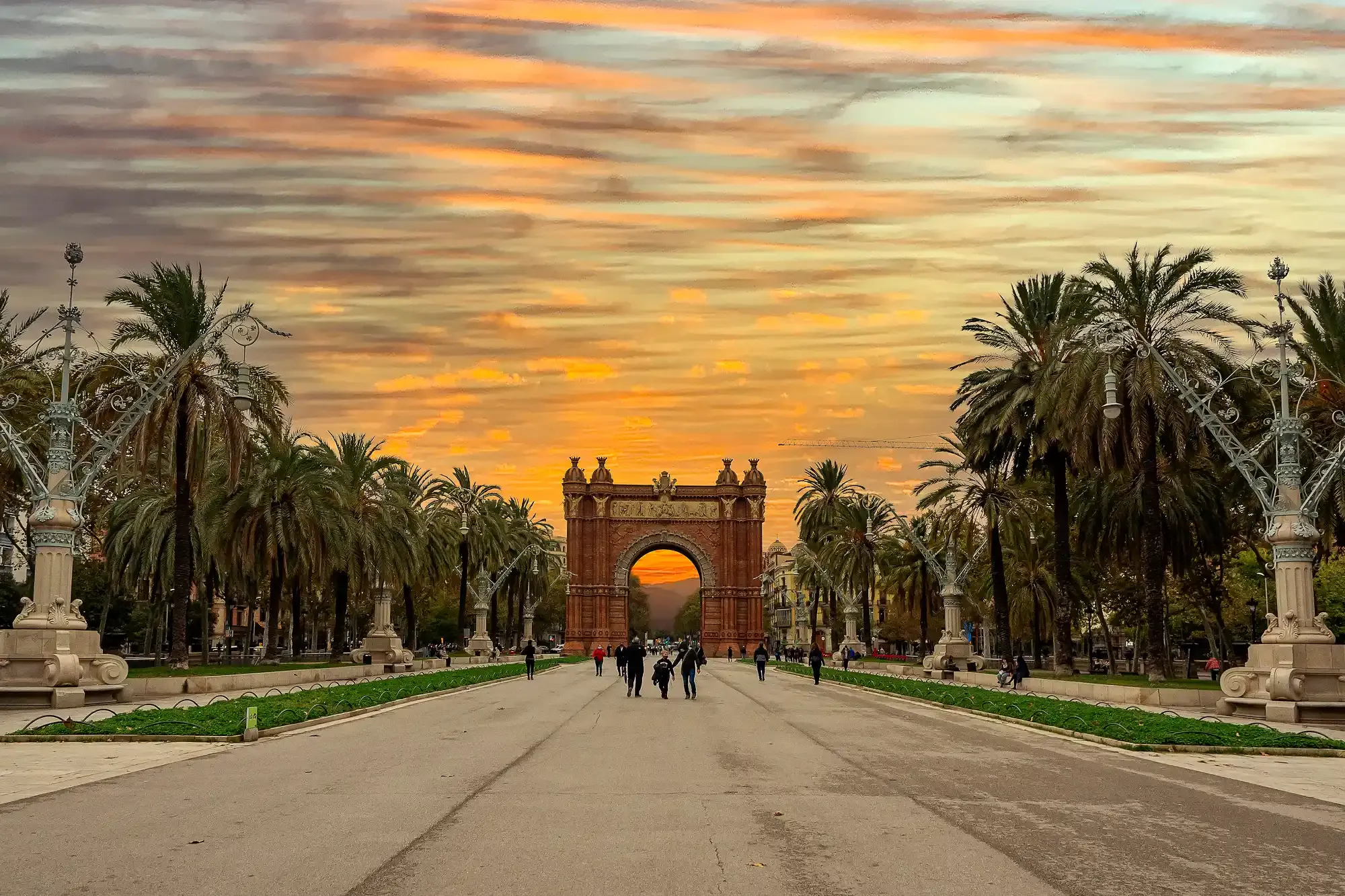 The Arc de Triomf in Barcelona at the end of a palm-lined promenade, under a vibrant orange sunset sky.