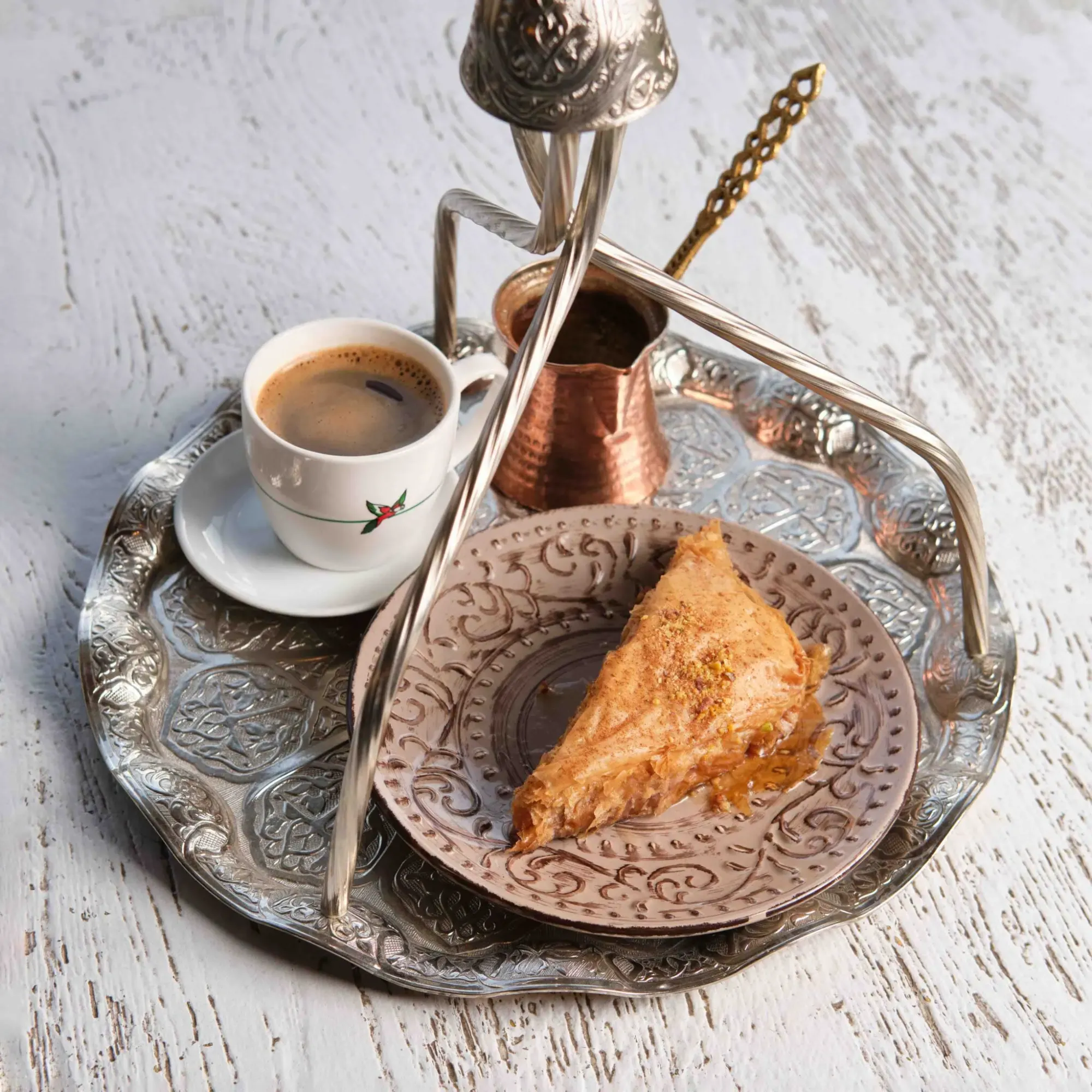 A silver tray holds a white cup of coffee, a copper cezve, a golden spoon, and a syrupy baklava piece on a patterned plate.