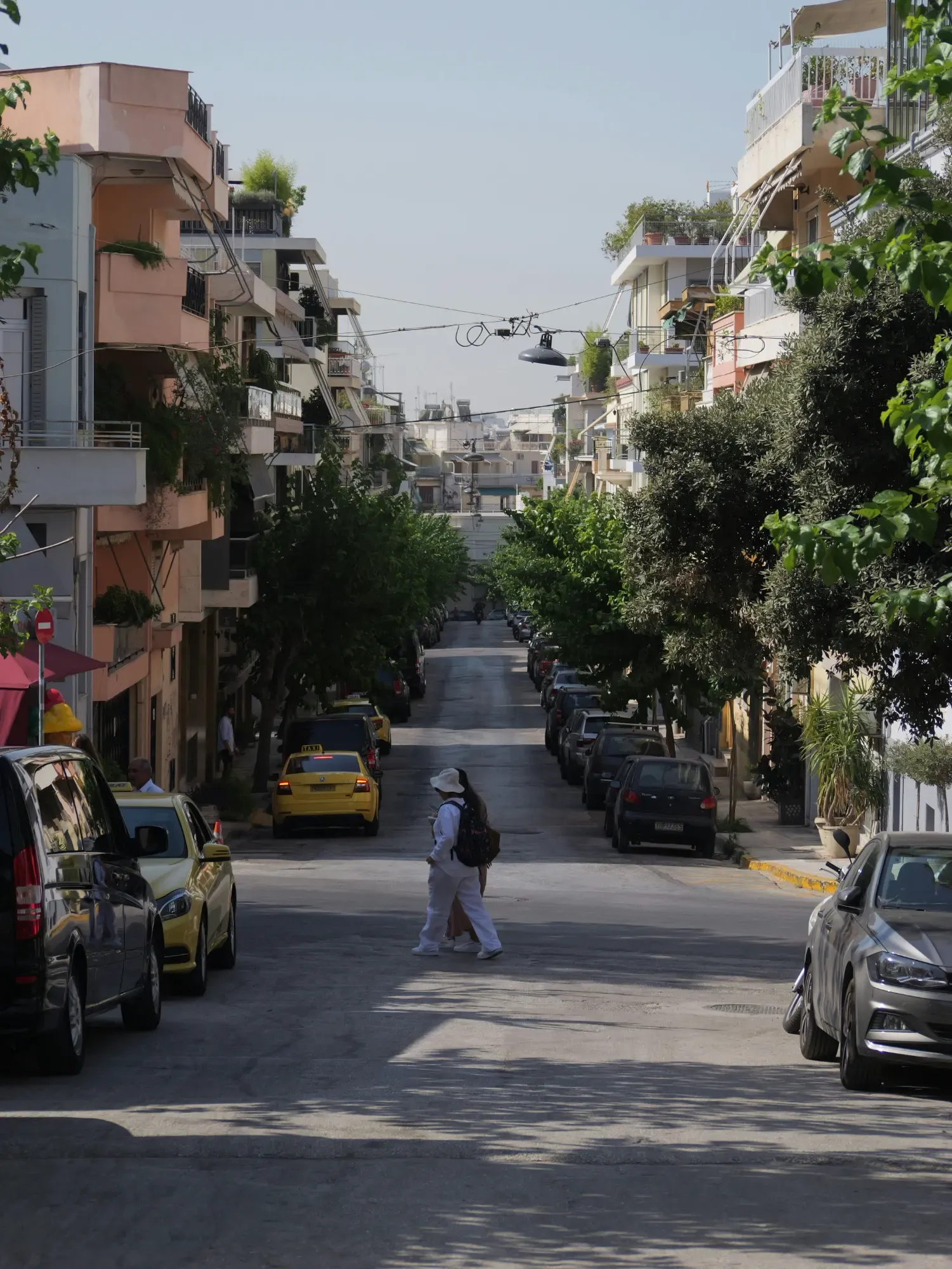 Residential street in Athens with a person walking down the center, flanked by parked cars and multi-story buildings with bal