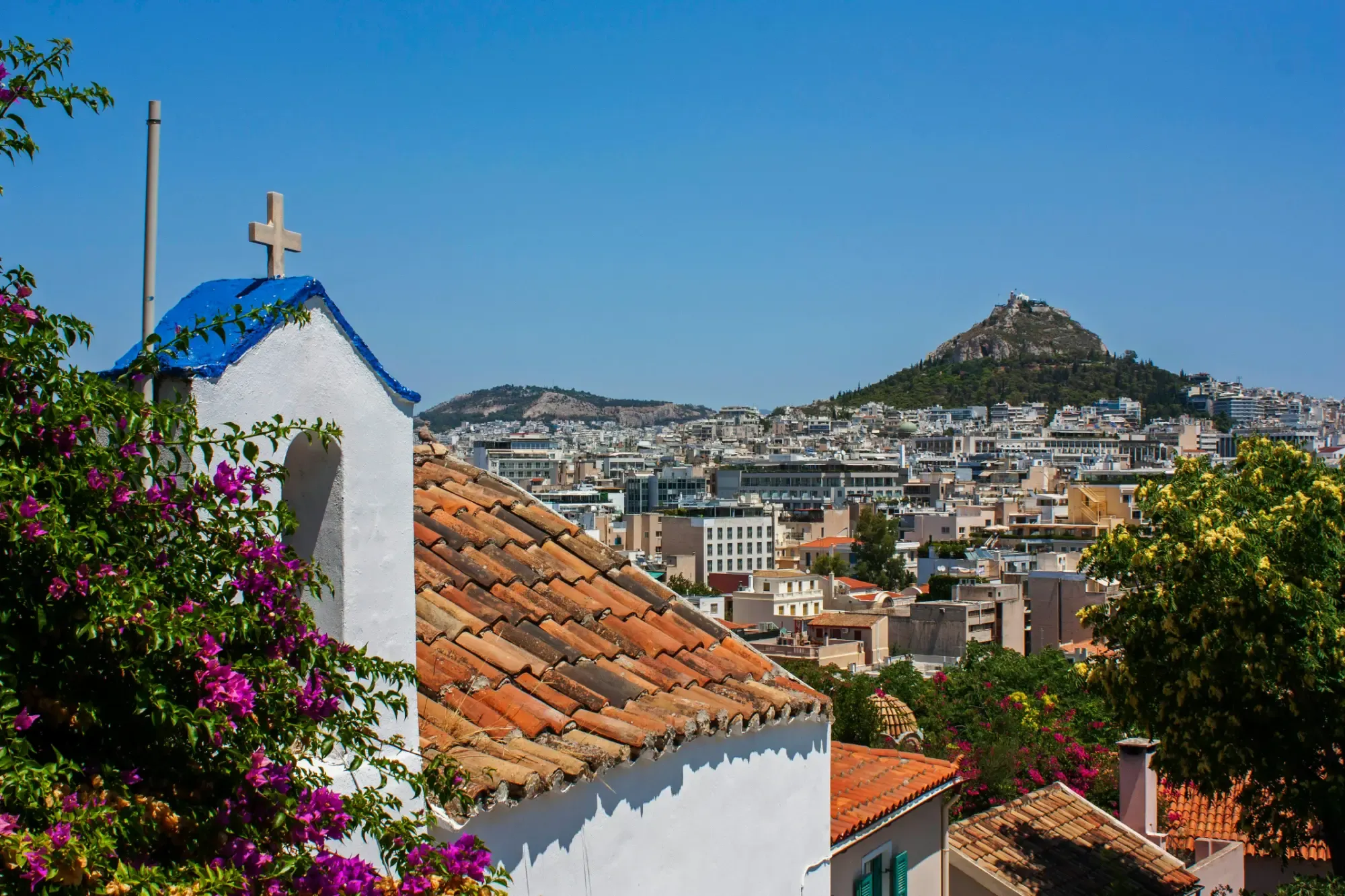 White chapel with blue roof and cross, bougainvillea, red tiled roofs, and Athens city under a clear sky with Lycabettus Hill
