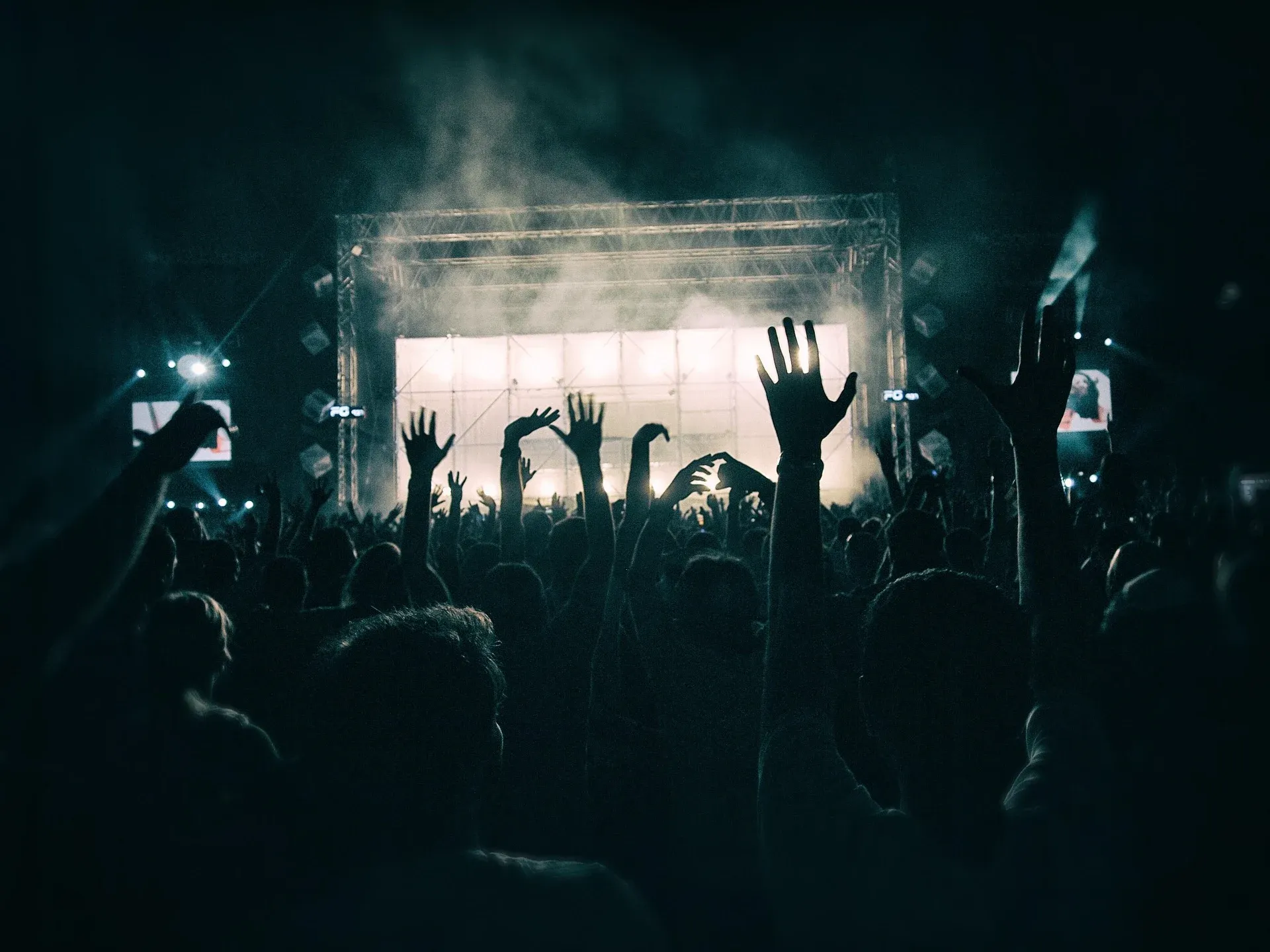 A crowd with hands raised at an outdoor concert in front of a bright stage at night