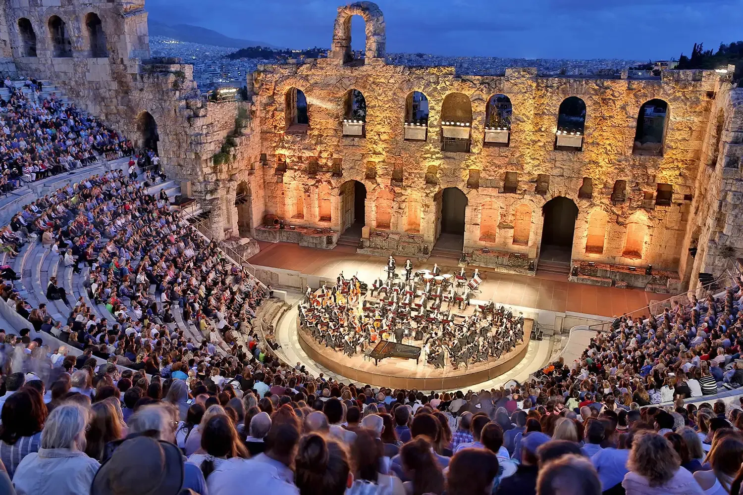 An orchestra performs for a large audience in the ancient stone amphitheater of the Odeon of Herodes Atticus at dusk.