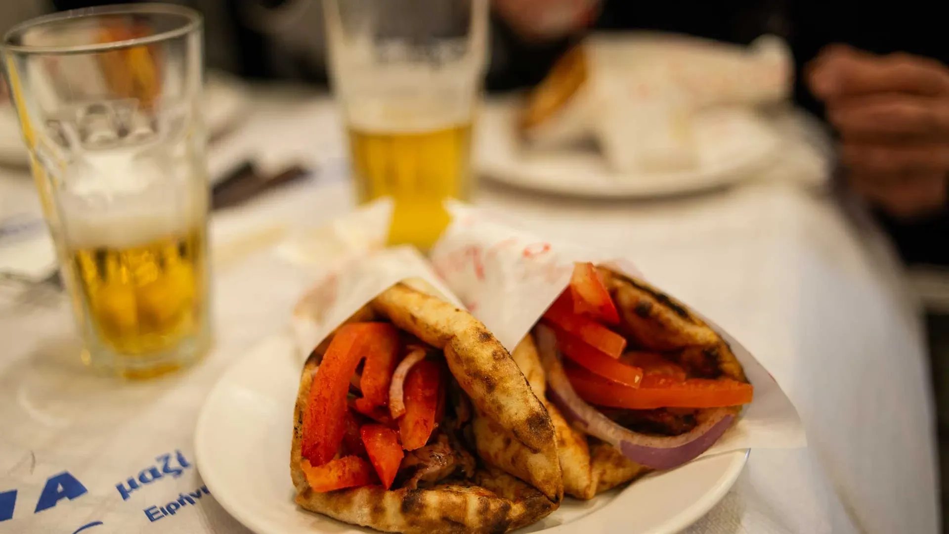 Zwei Souvlaki in Pitabrot auf einem weißen Teller, serviert mit Tomaten und Zwiebeln, dazu ein Glas Bier.