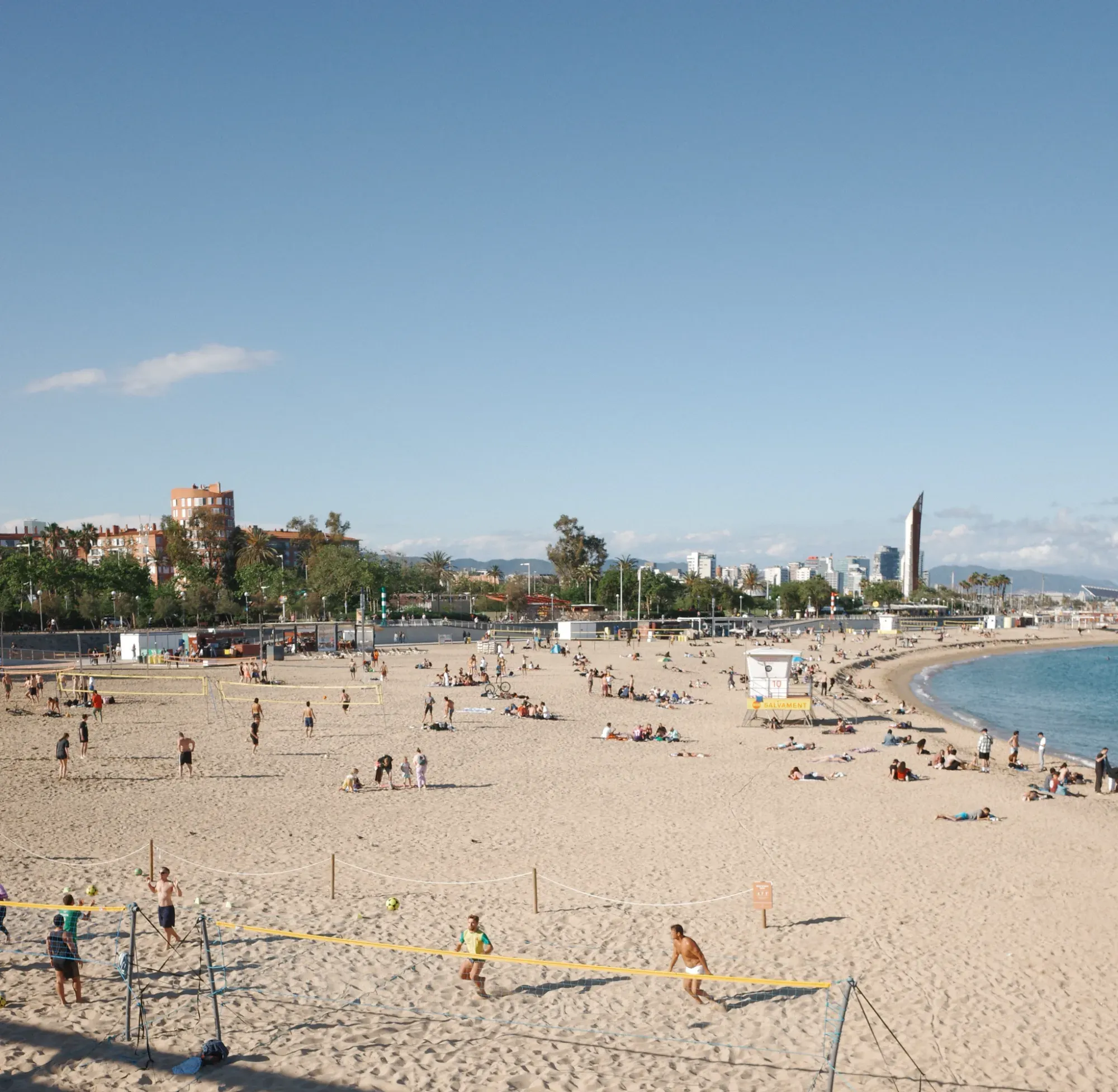 Un día soleado en una concurrida playa de Barcelona, con gente jugando al voleibol y relajándose en la arena.