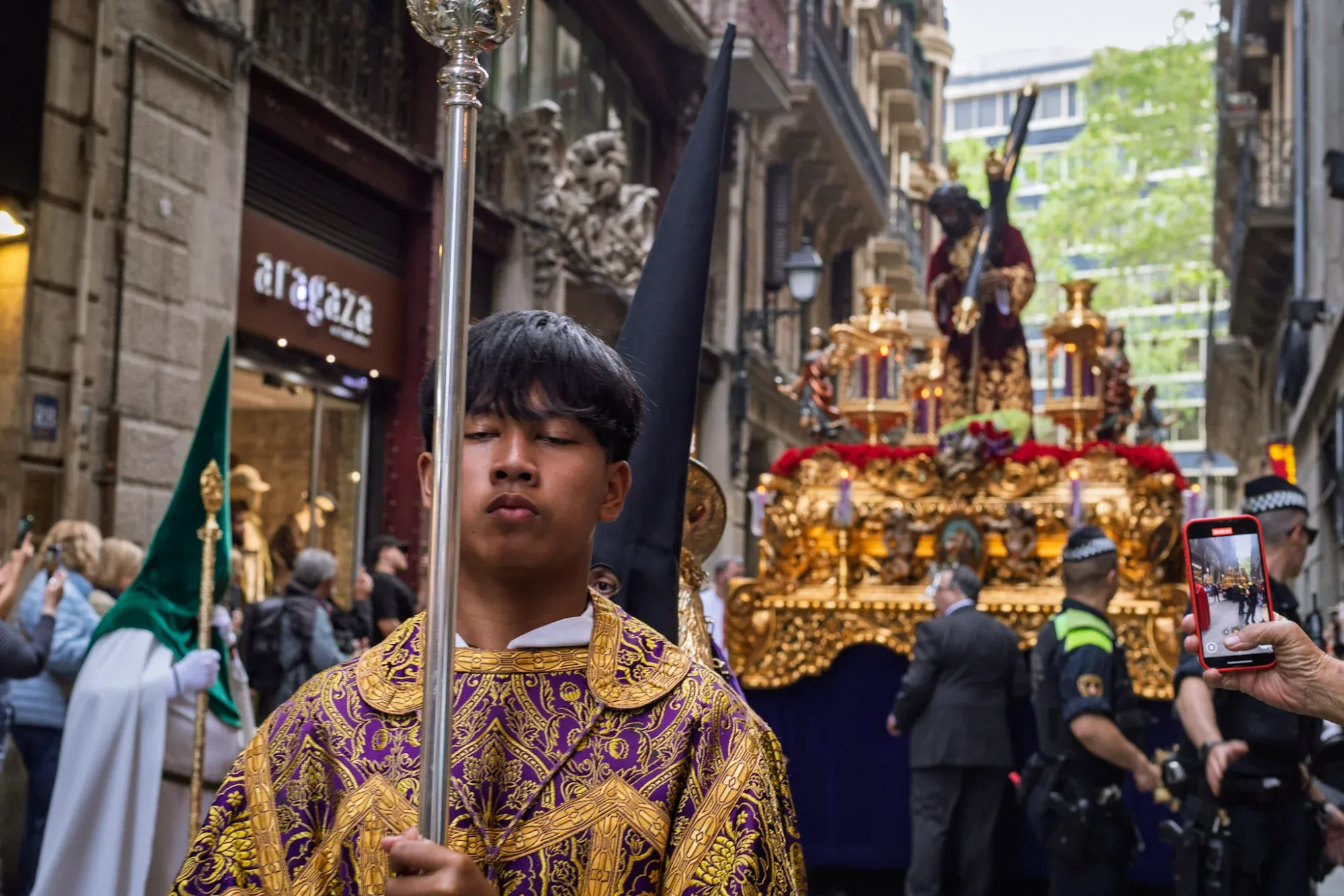 Person in purple and gold tunic with a staff, leading a Holy Week procession with a religious float and Jesus statue on a cit