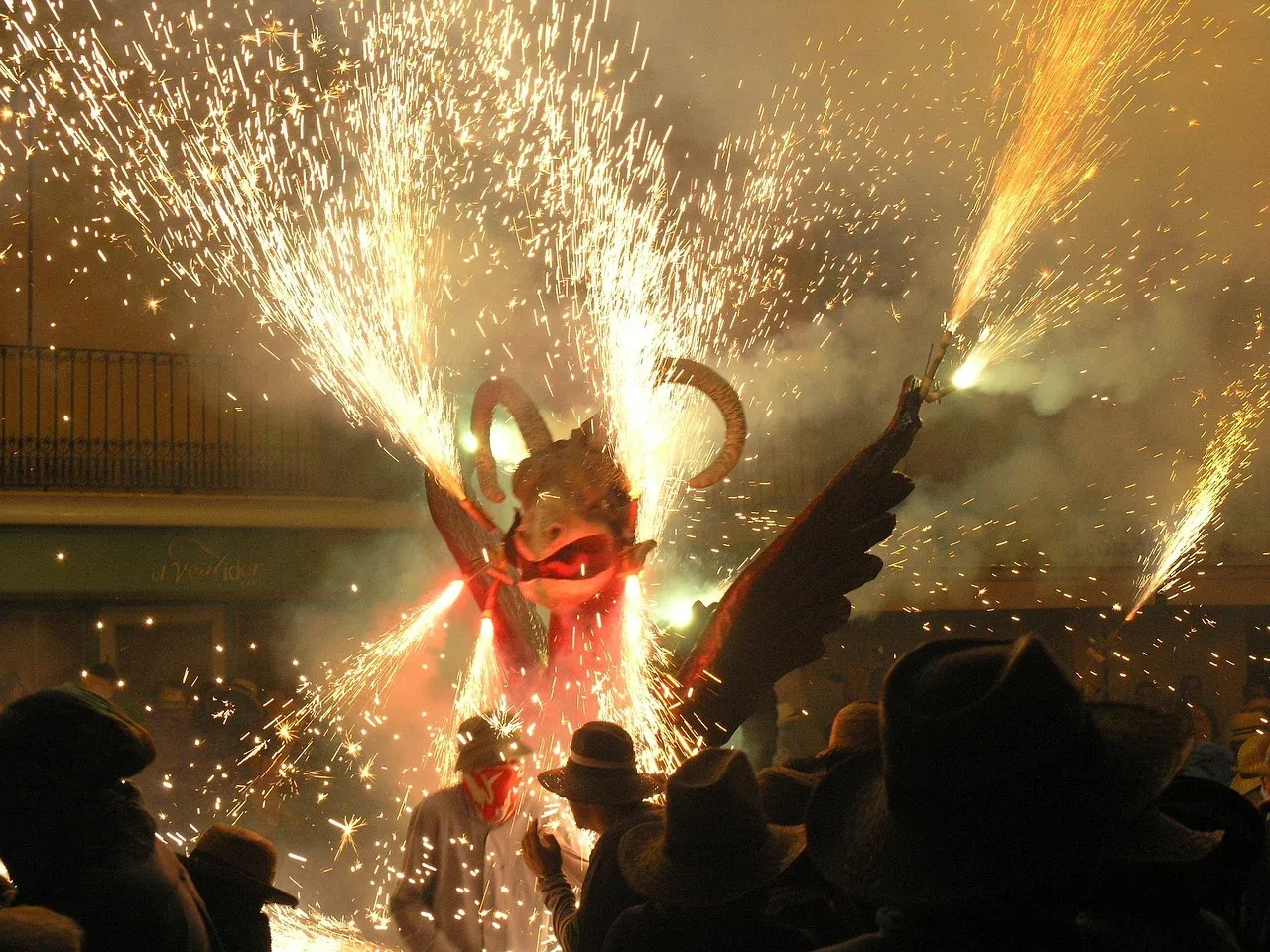 Large winged dragon figure with horns emitting sparks and fireworks at a night festival, watched by silhouetted people.