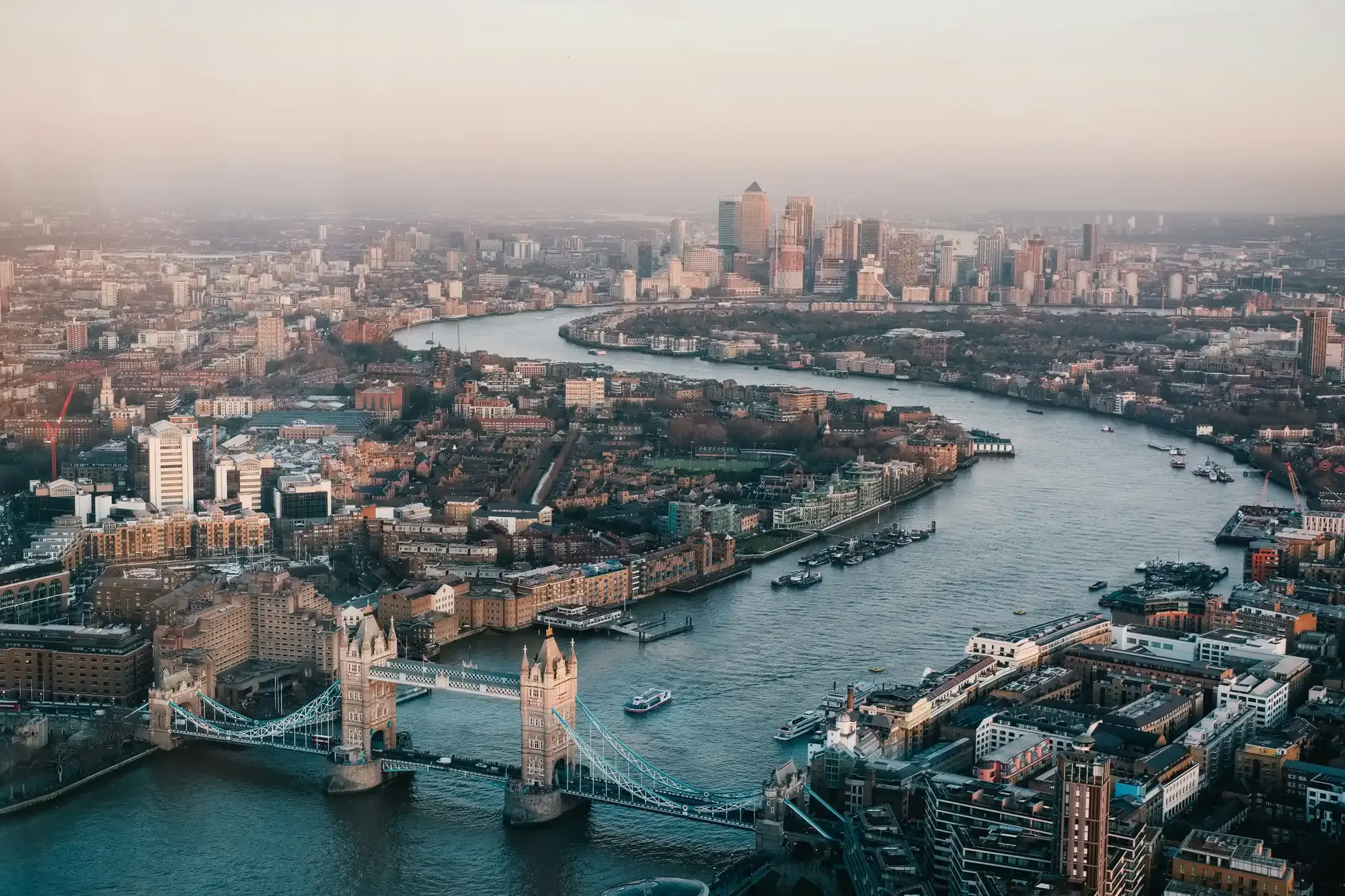 Vista aérea de Londres con el Puente de la Torre y el río Támesis, y el skyline de Canary Wharf a lo lejos al atardecer.