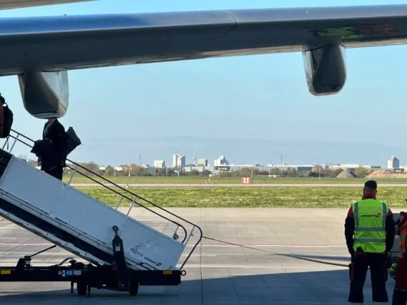 Parte inferior del ala de un avión con pasajeros abordando por escaleras móviles, personal de tierra del aeropuerto y una sil
