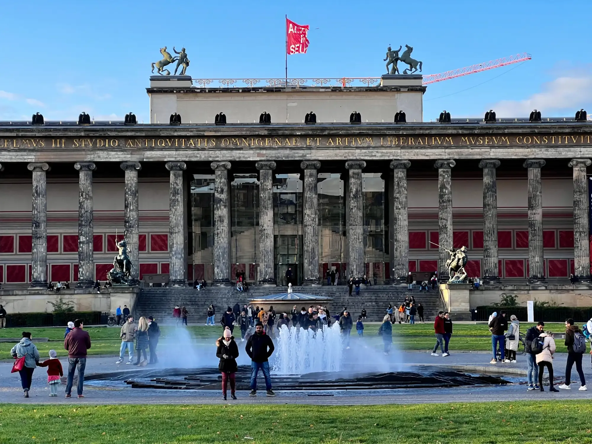 Klassische Fassade des Alten Museums mit Säulen, Dachstatuen, roter Flagge und einem Brunnen im Lustgarten, mit Menschen.