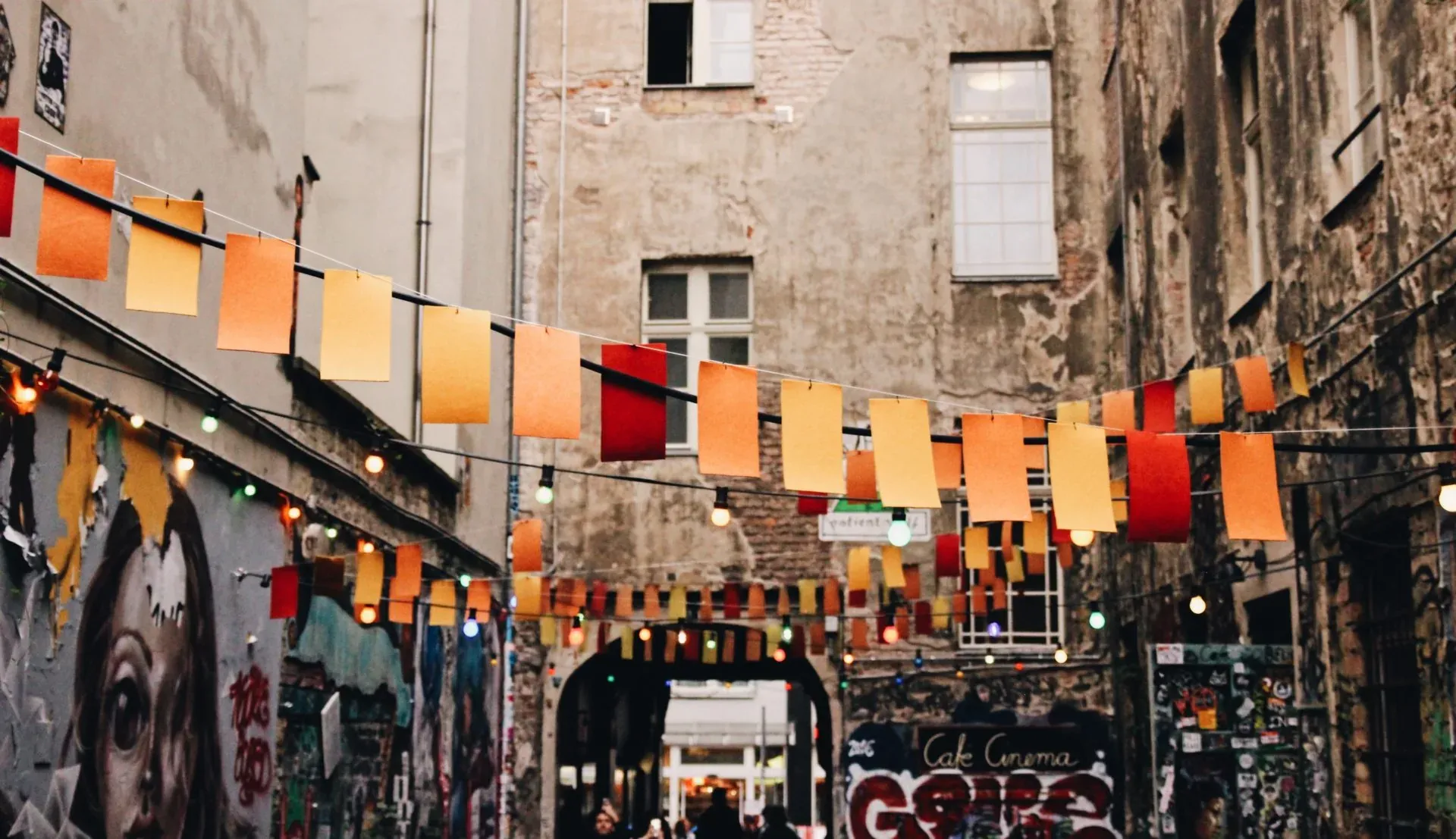 An alley in Berlin with street art and colorful flags and lights strung between weathered apartment buildings.