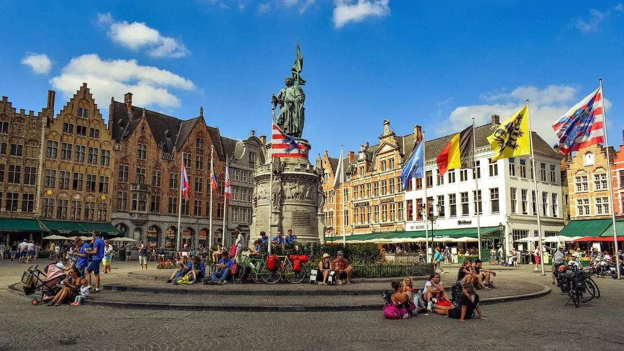 Large public square in Bruges with a central statue on a pedestal, surrounded by historic buildings and people sitting or wal