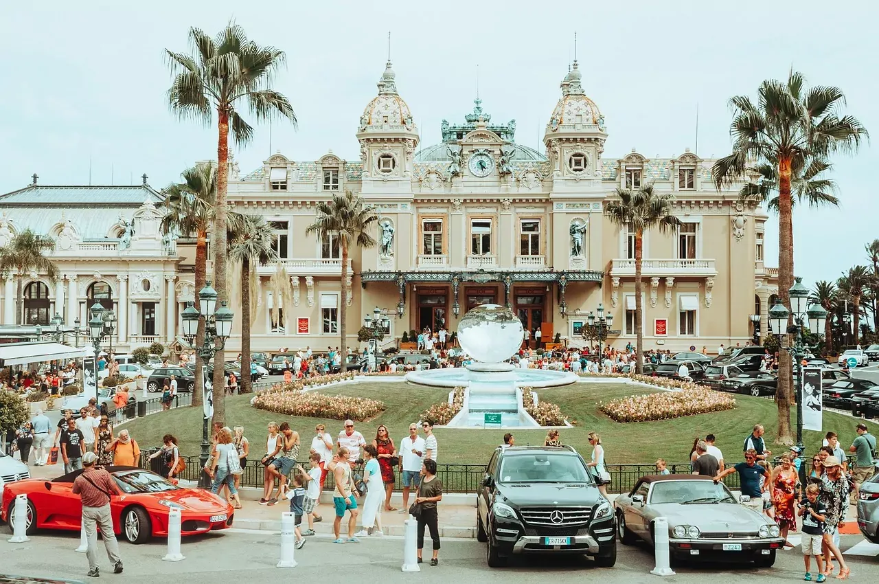 Grand facade of Monte Carlo Casino with palm trees, globe fountain, luxury cars, and many visitors on a clear day.