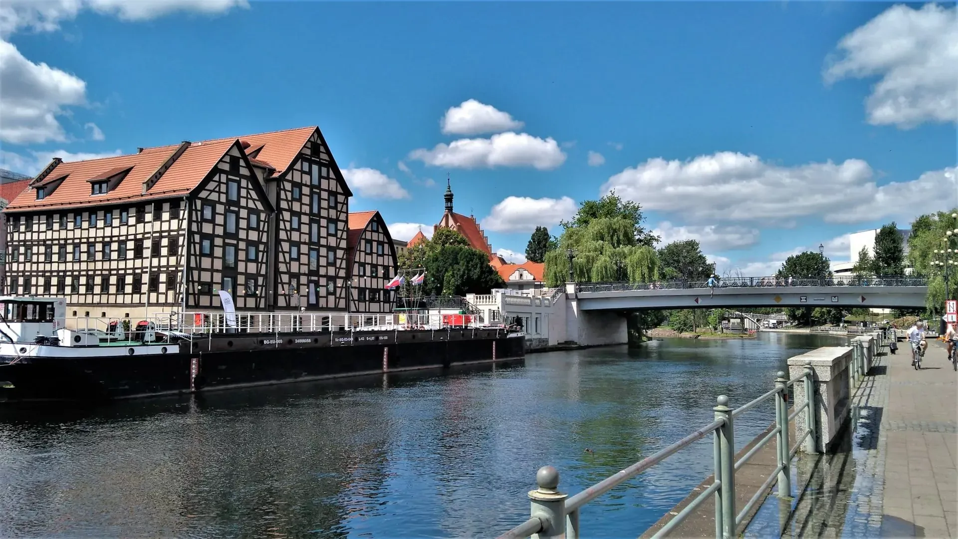 Spichrze nad rzeką Brdą, most, barka i piesi na słoneczny dzień.