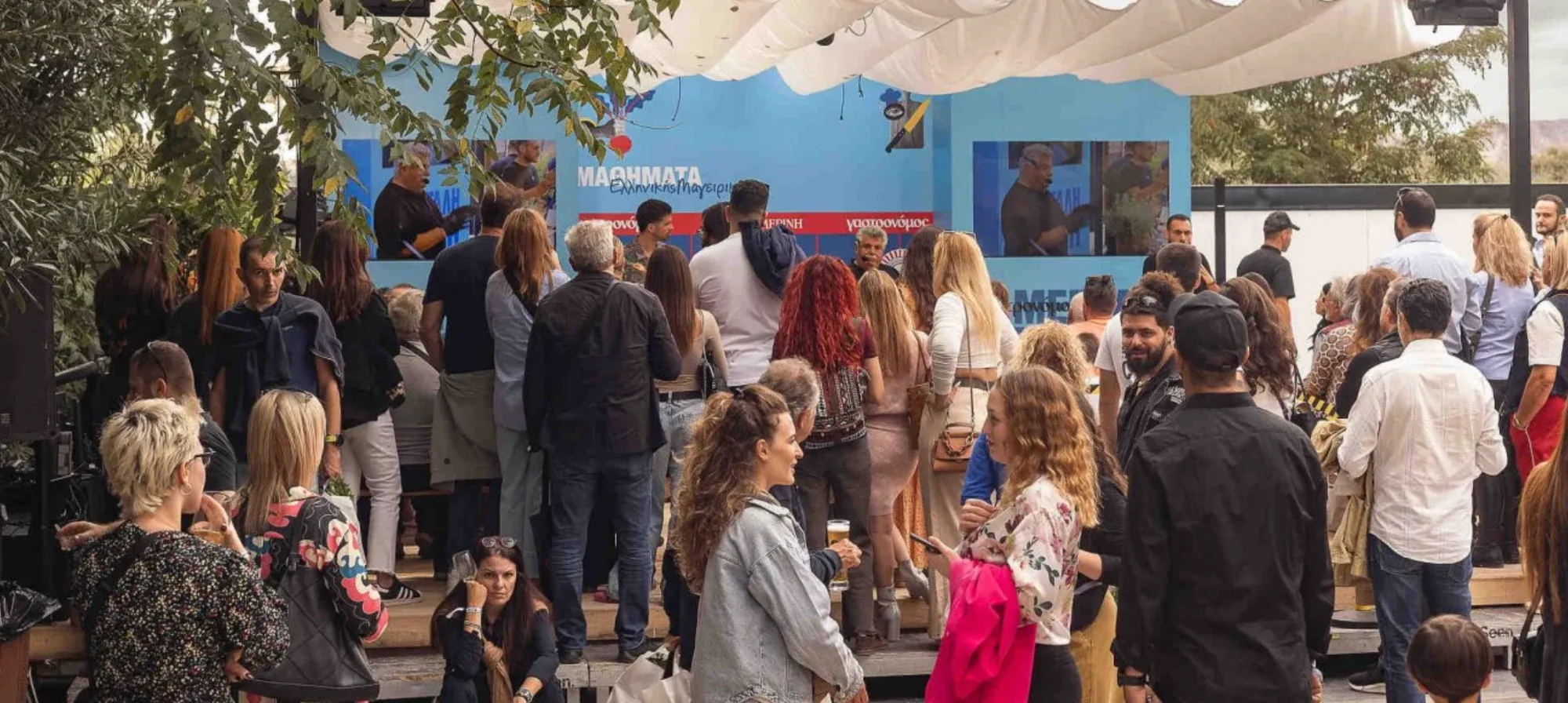A crowd of people gathered outdoors at an event, watching a presentation on a blue stage.
