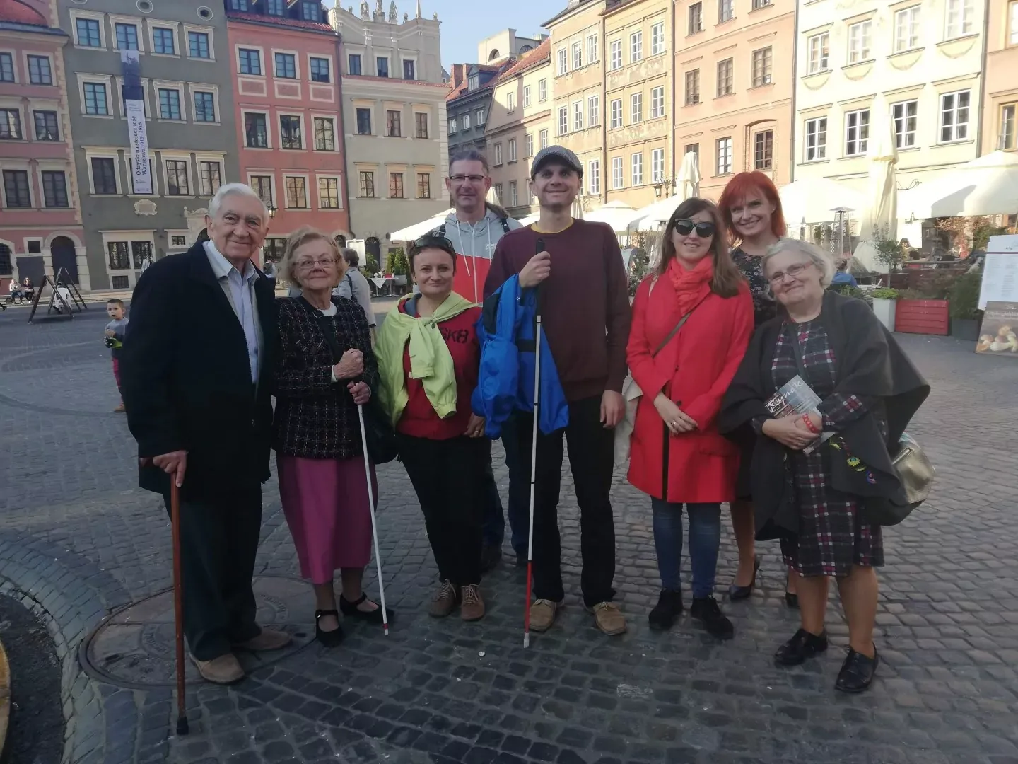 Eine vielfältige Gruppe von Menschen, darunter einige mit Blindenstöcken und einem Gehstock, steht auf einem Kopfsteinpflaste