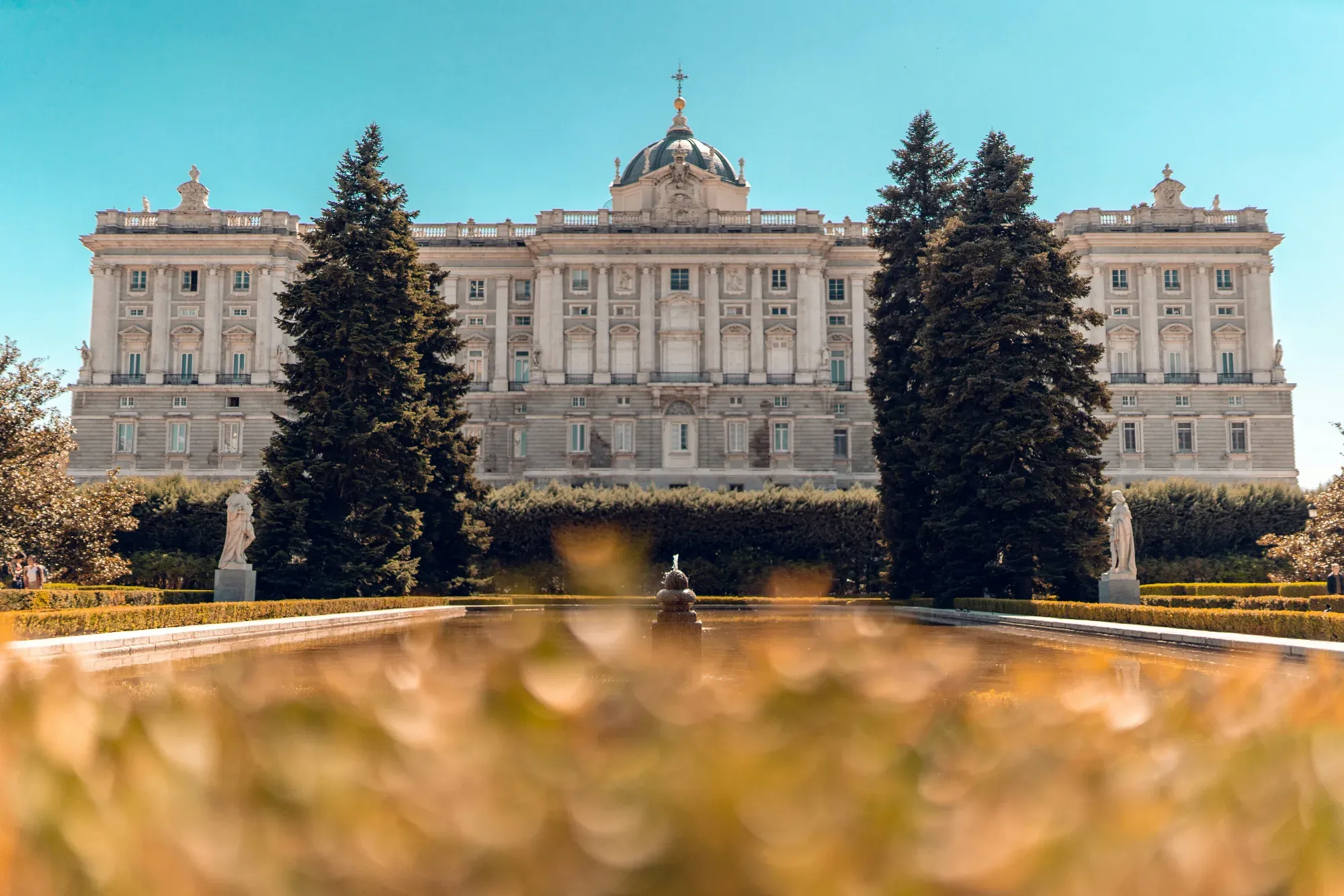 Royal Palace of Madrid seen across a pond in Sabatini Gardens flanked by evergreen trees and statues.