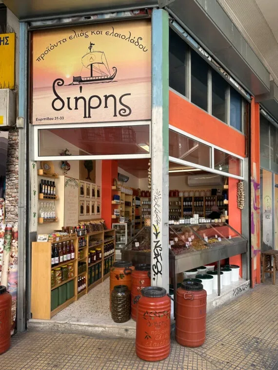 Straßenansicht eines Athener Ladens für Olivenprodukte und Öl, mit roten Fässern draußen und Regalen voller Gläser drinnen.