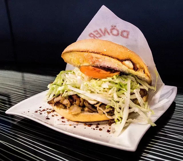 Un Döner Kebab en pan con carne, col rallada y una rodaja de tomate, servido en un plato blanco. Envoltorio de papel con el t