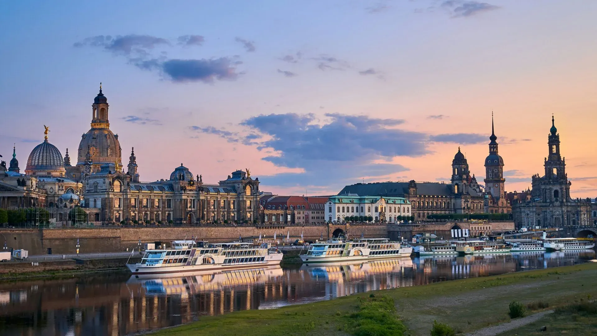 Dresden skyline at sunset with Frauenkirche dome, Hofkirche, and boats on the Elbe River reflecting colors.