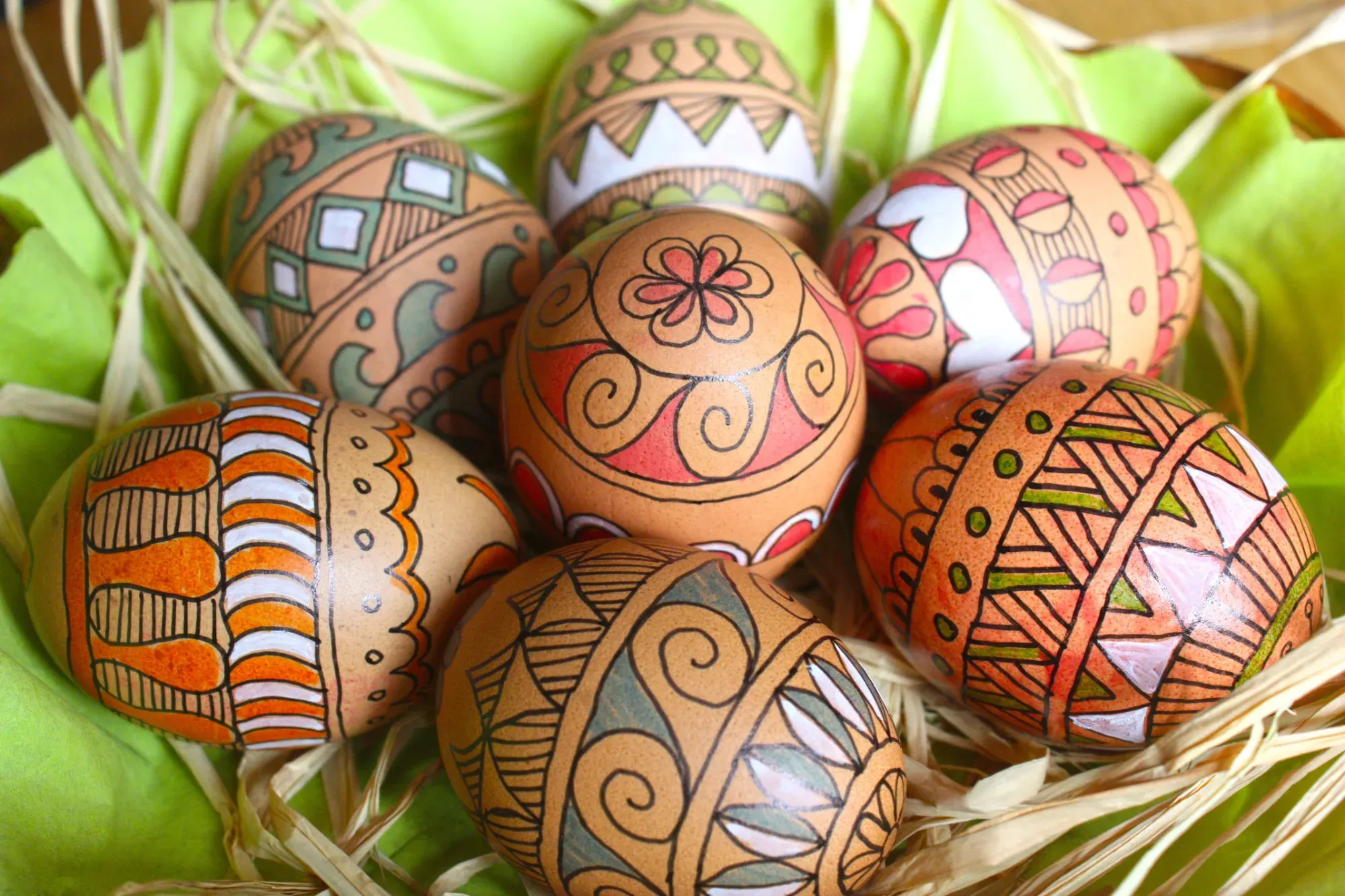 Sieben braune Ostereier mit detaillierten rot, grün, orange und weißen Mustern, in einem Nest aus Stroh und grünem Stoff.
