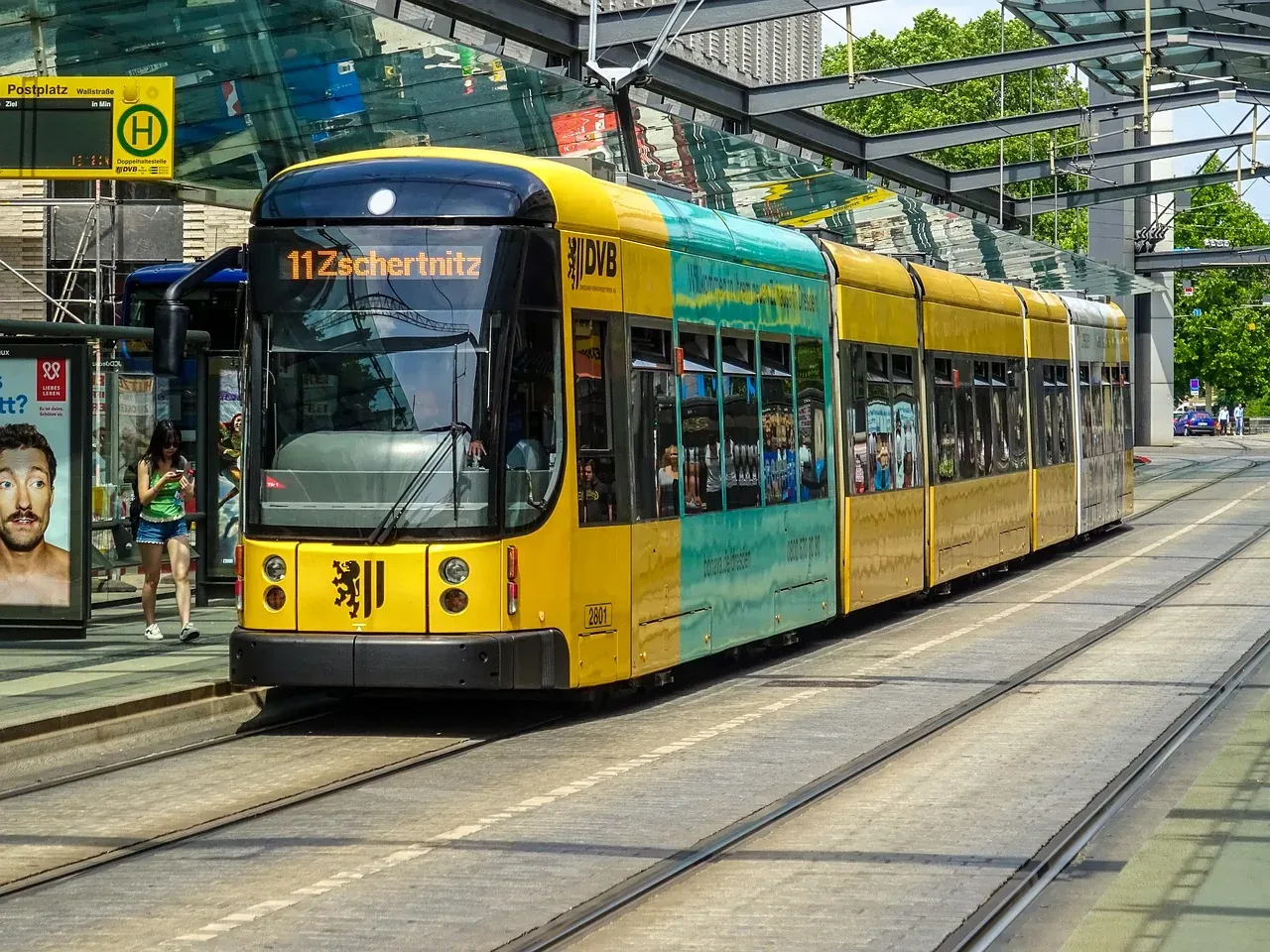 A yellow and teal Dresden tram, line 11 Zschernitz, on tracks at a city stop with a glass shelter. A woman stands nearby.