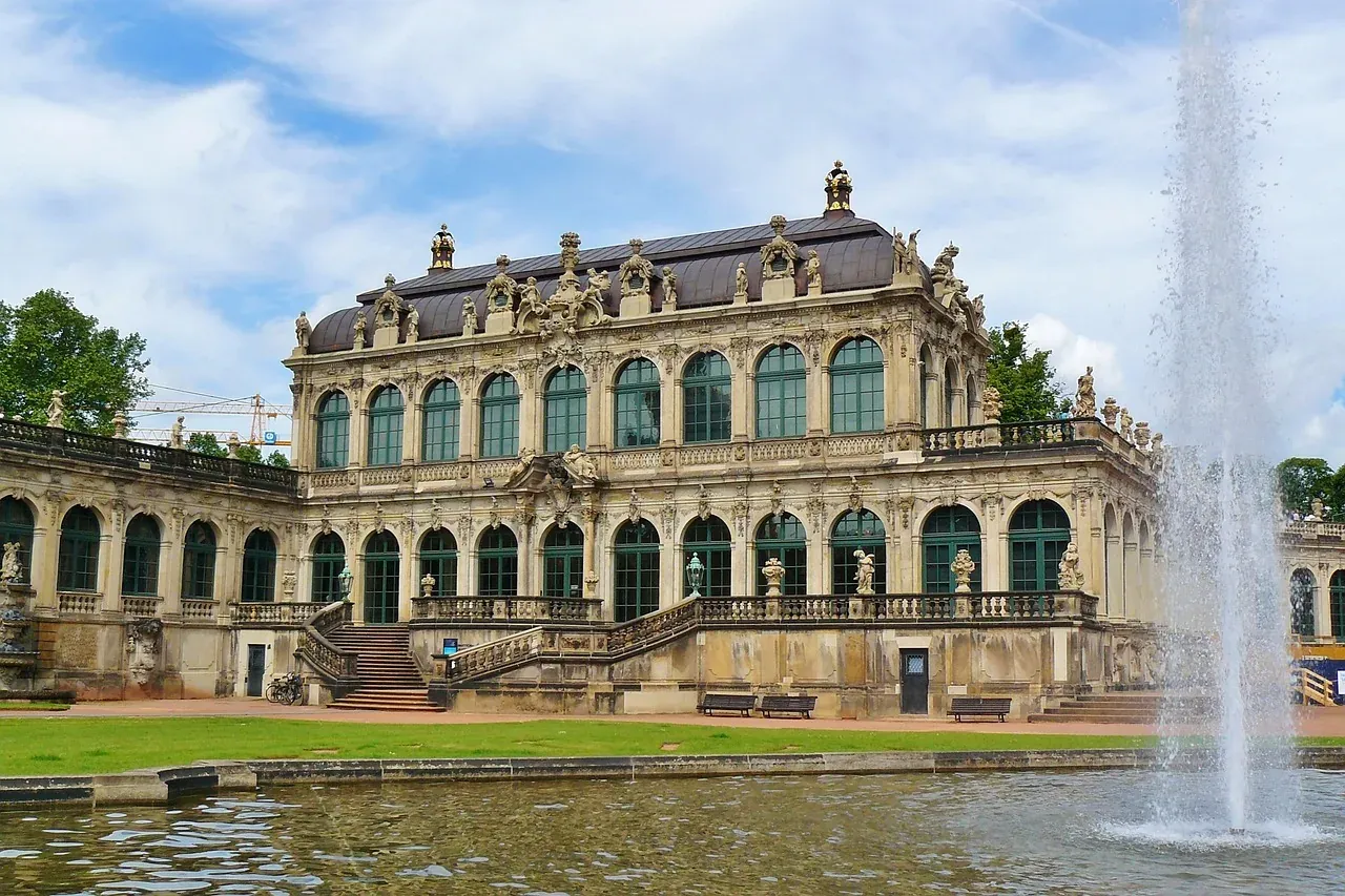 Baroque palace facade with green-framed windows and ornate details, seen across a pond with a large fountain spraying water.