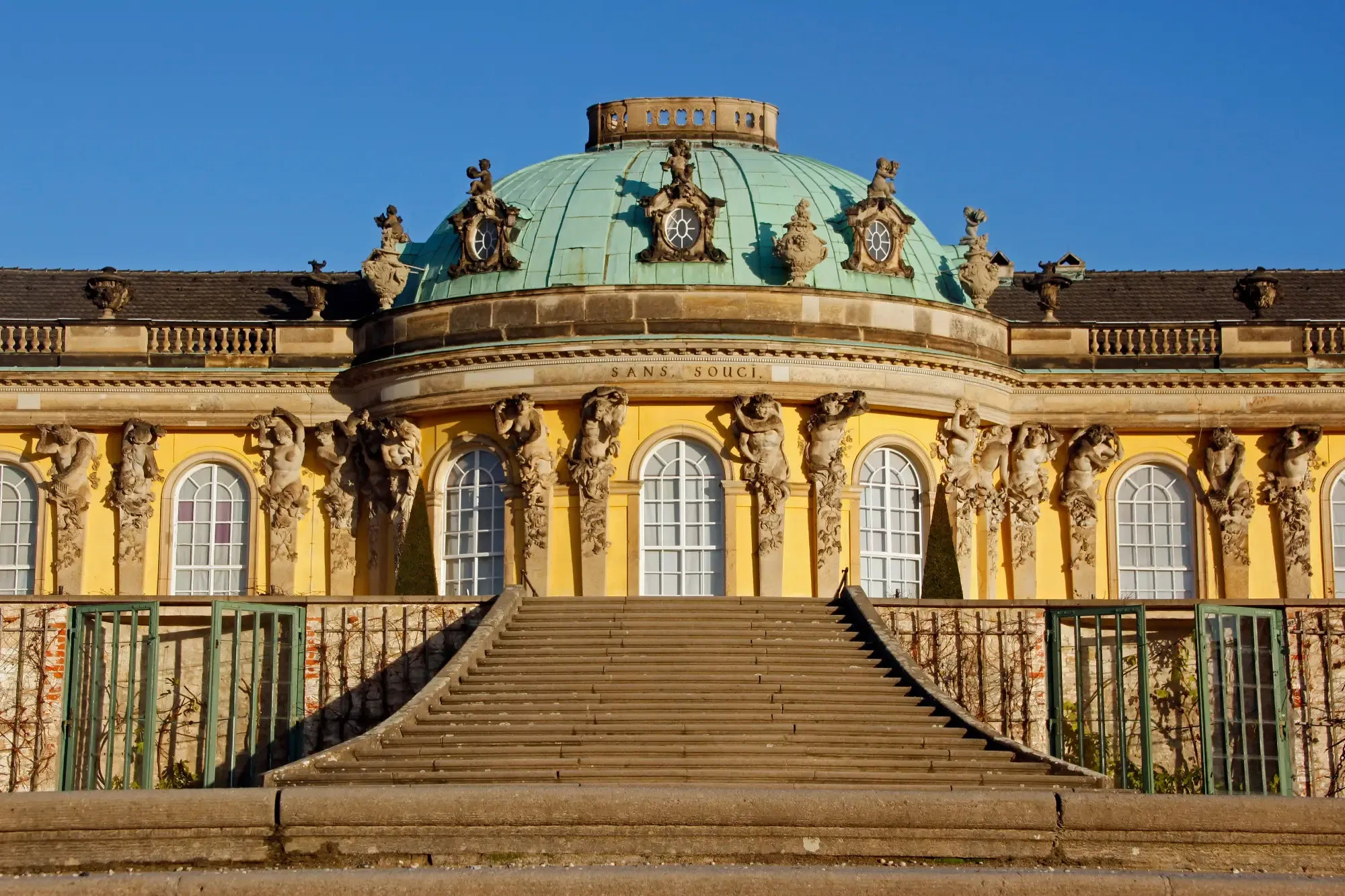 Fachada amarilla y cúpula verde del Palacio de Sanssouci en Potsdam, con escalinatas de piedra.