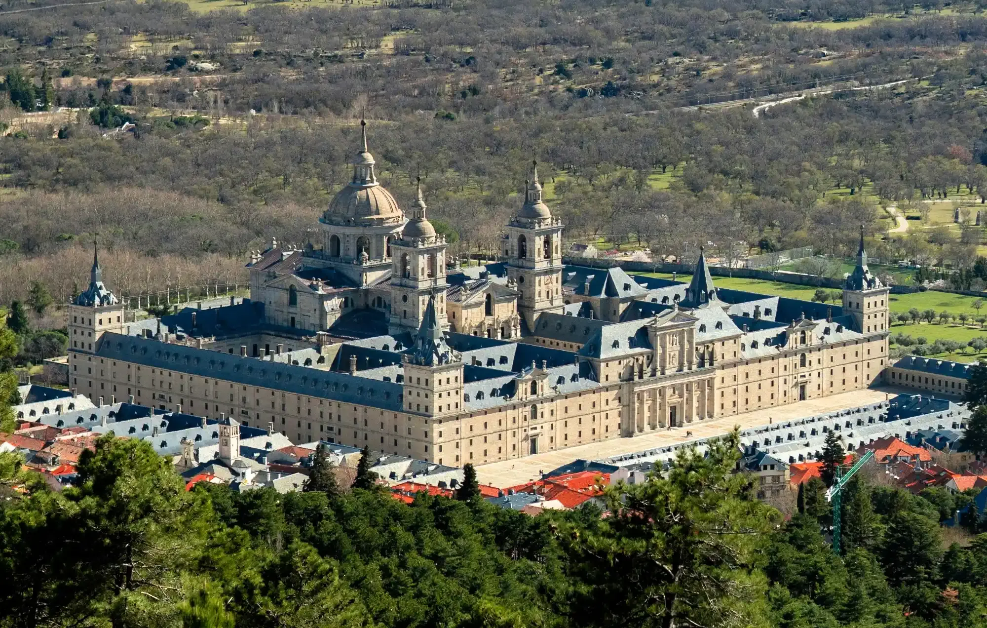 Aerial view of the San Lorenzo de El Escorial Royal Site, a large stone monastery complex with domes and spires, surrounded b