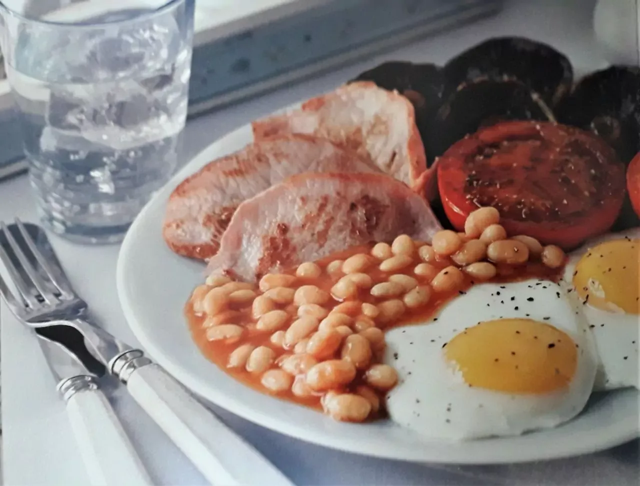A plate with two fried eggs, baked beans, grilled bacon, a grilled tomato, and black pudding. A glass of water and cutlery ar