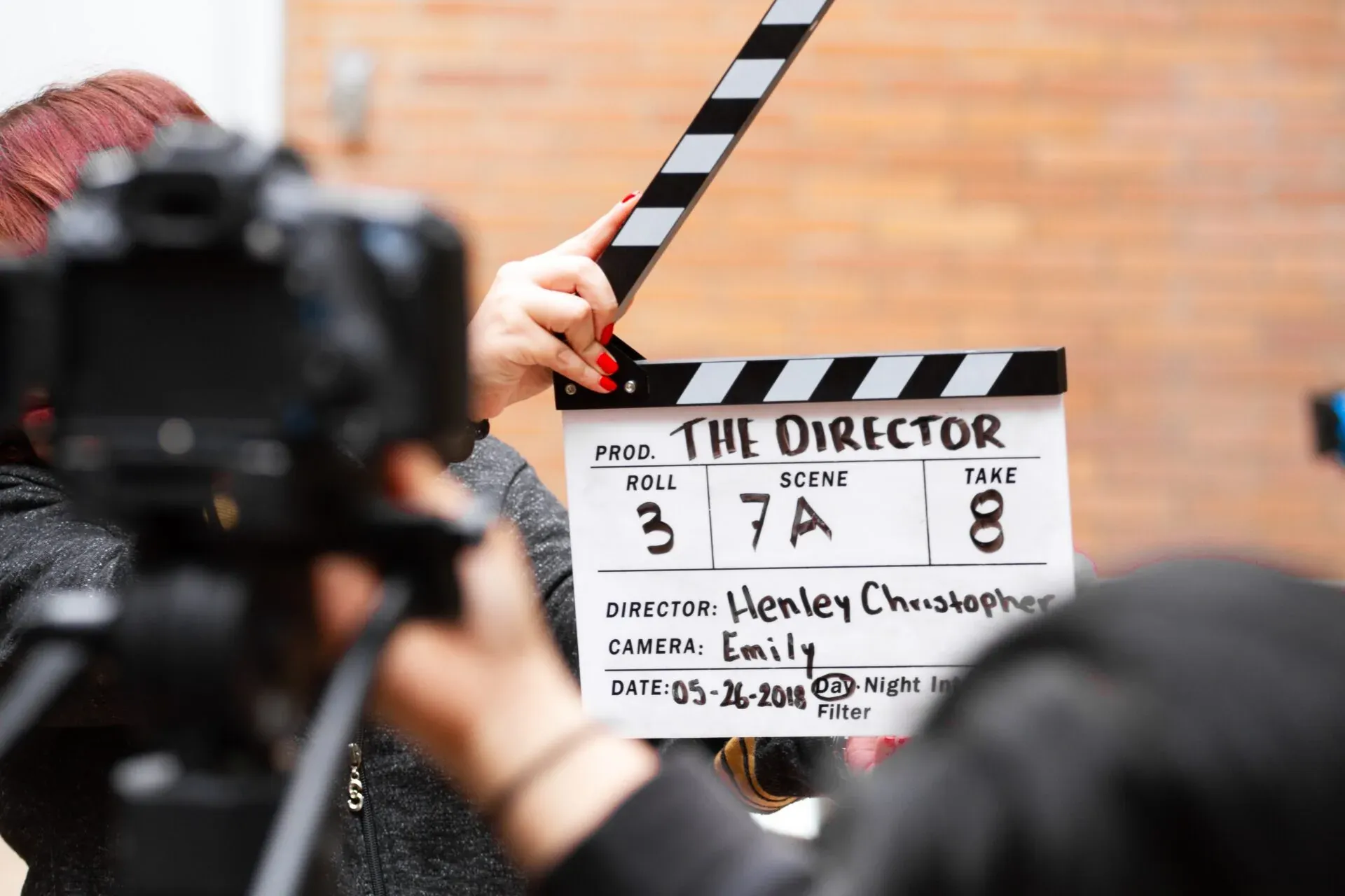 Close-up of a clapperboard with production details and a hand with red nail polish holding it, camera in foreground.