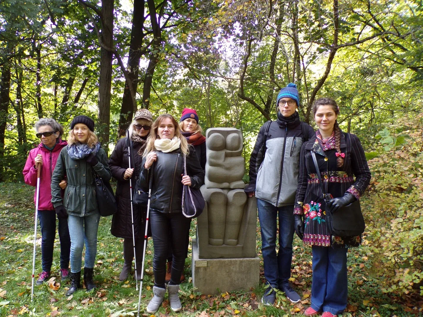 Group of seven people, some with white canes, standing in a park next to a stylized stone sculpture. Green and autumn foliage