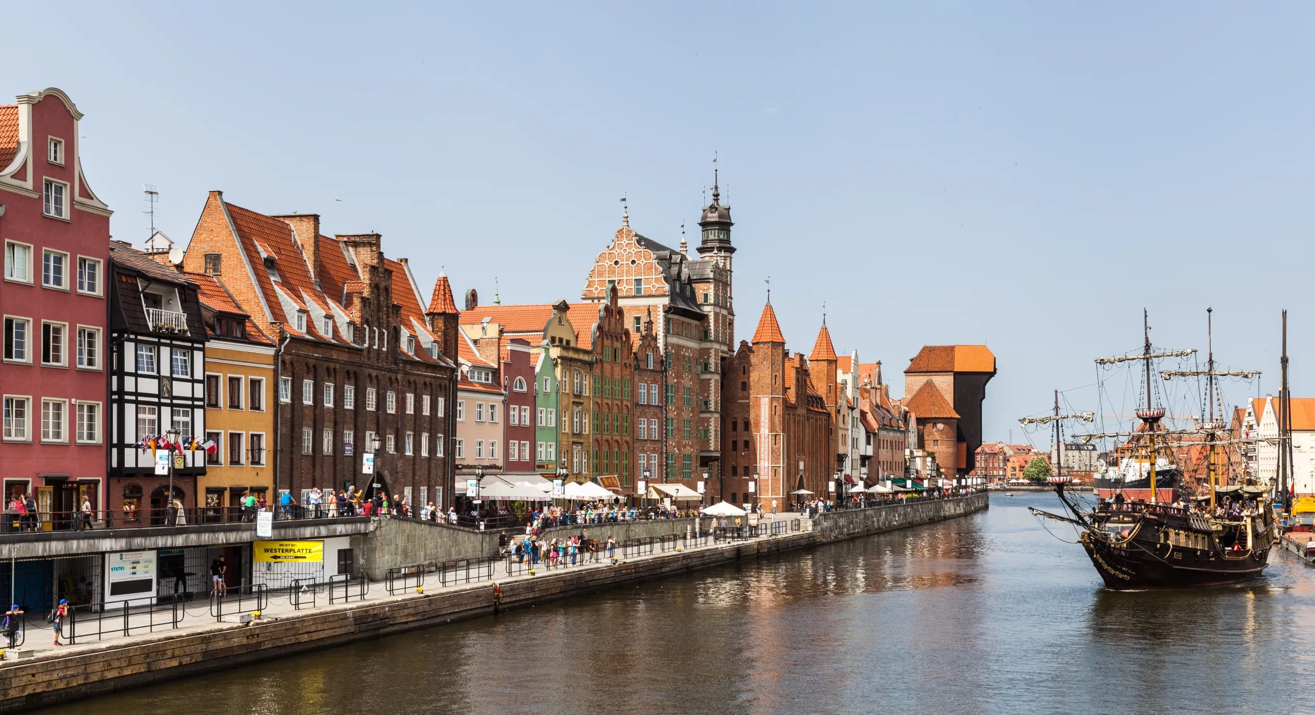 Historyczne kamienice wzdłuż rzeki Motławy w Gdańsku, z ludźmi na promenadzie i dużym statkiem. Widoczny jest Żuraw.