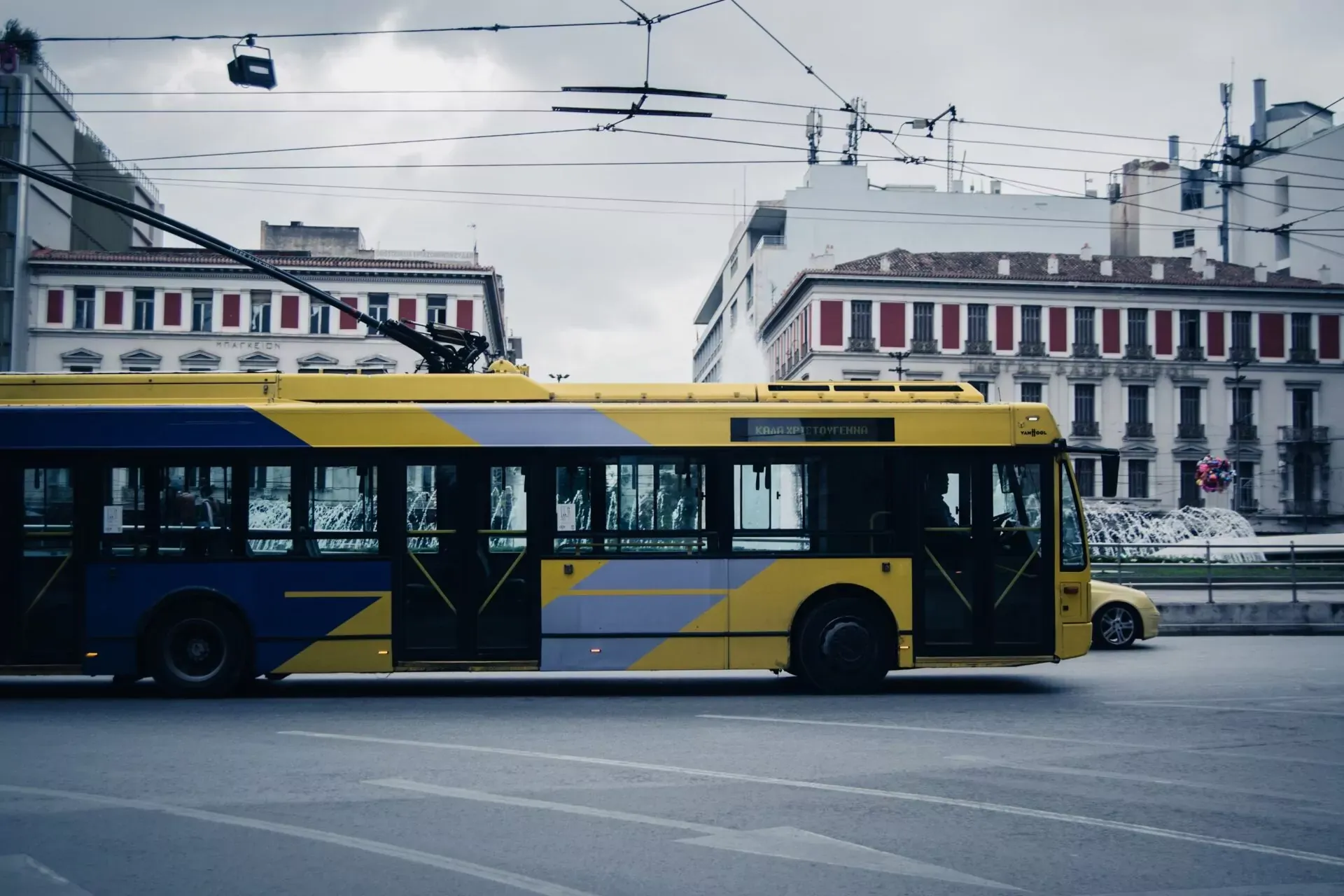 Żółto-niebieski trolejbus elektryczny w ruchu na ulicy w Atenach, z budynkami i liniami trakcyjnymi na tle zachmurzonego nieb
