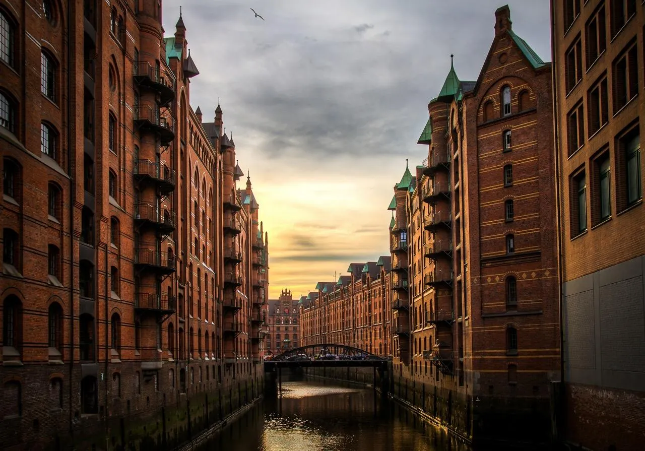 Historic brick warehouses of Speicherstadt in Hamburg lining a canal at sunset, with warm light and a bridge.