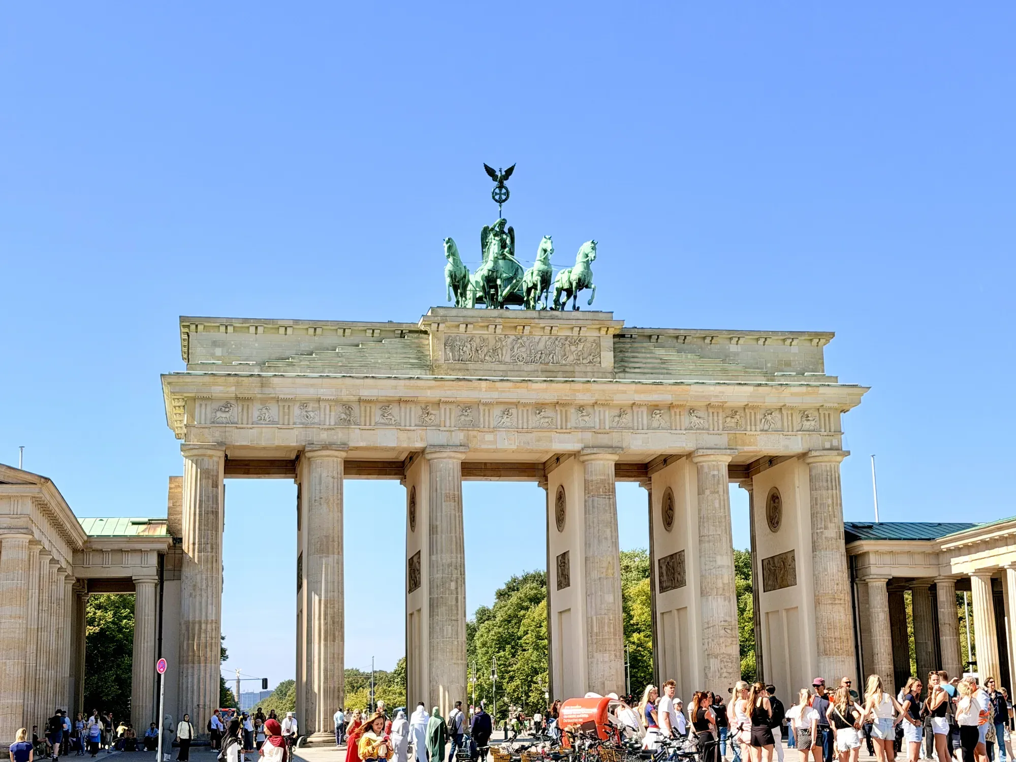The Brandenburg Gate in Berlin on a sunny day, with the Quadriga statue on top and crowds of tourists below.