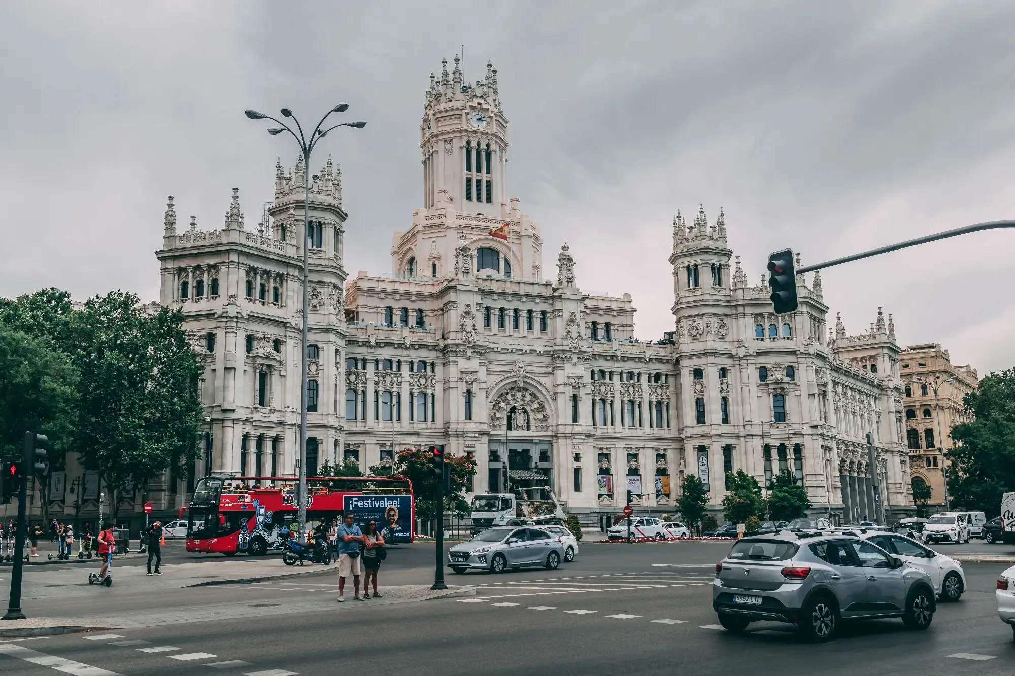 Die prächtige weiße Fassade des Palacio de Cibeles in Madrid, davor eine belebte Straßenszene mit Autos und einem Tourbus.
