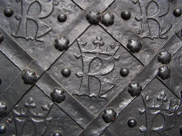 Close-up of an old, dark metal door with an embossed 'R' and crown design, set within a studded grid pattern.