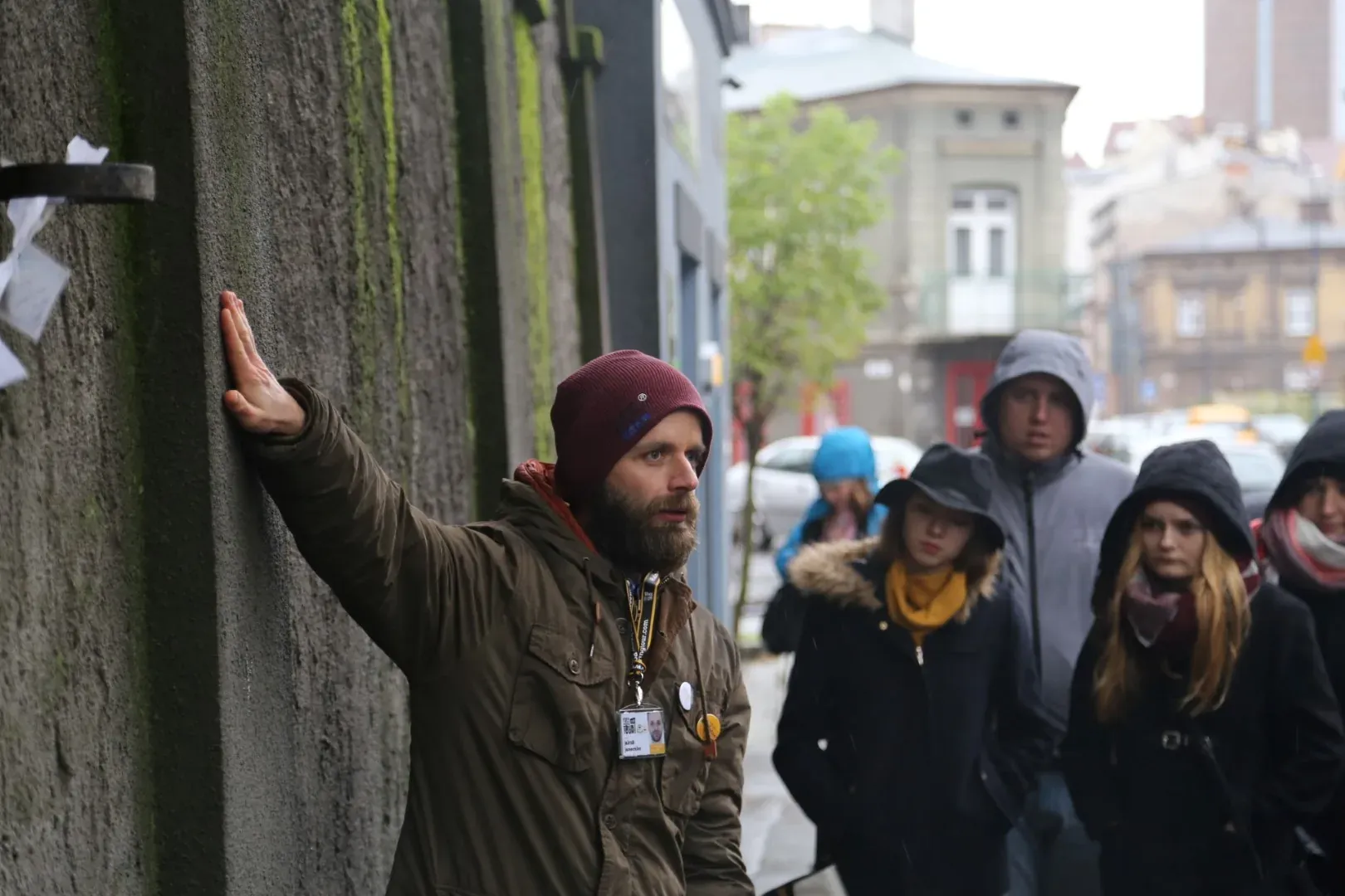 Guía barbudo con gorro burdeos toca una pared de hormigón musgosa mientras lidera un grupo en la calle.