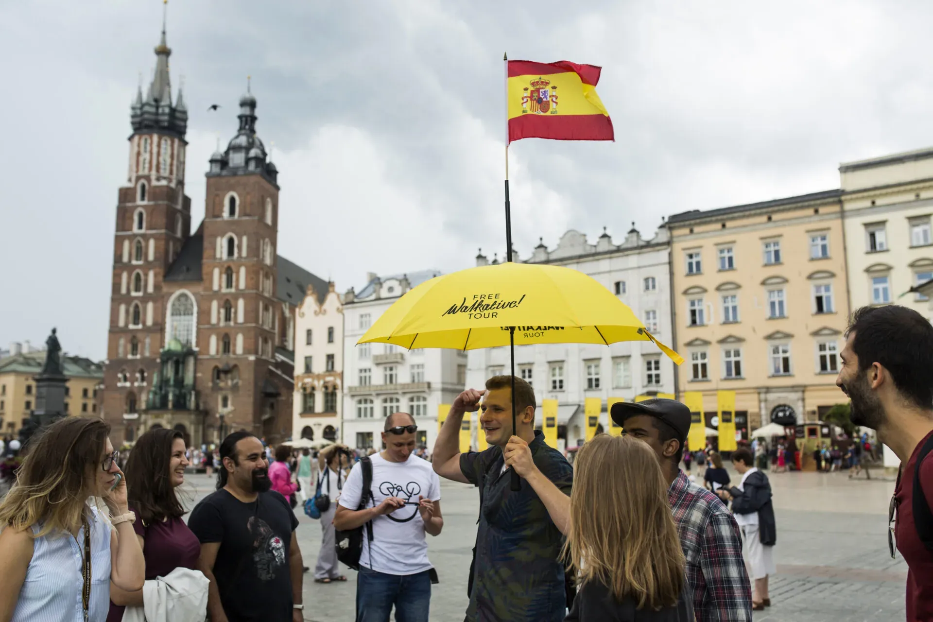 Group of tourists on a walking tour in Kraków's Main Market Square. A guide holds a yellow Walkative! umbrella with a Spanish