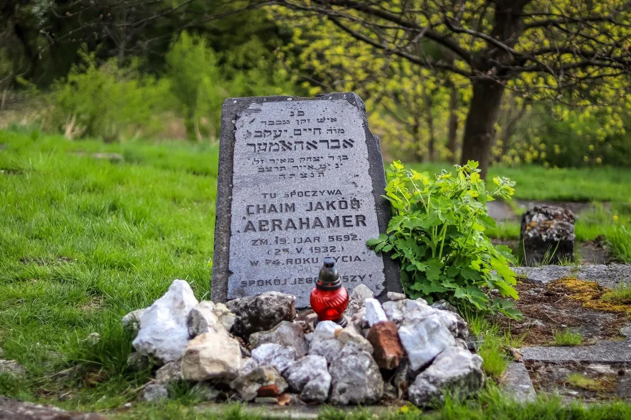 Verwitterter grauer Grabstein mit hebräischen und polnischen Inschriften, einer roten Gedenkkerze und Steinen auf Gras.